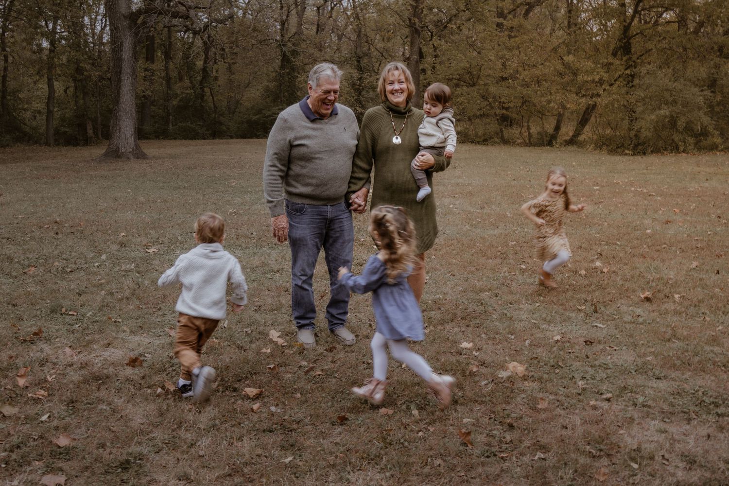 Grandparents playing outdoors in autumn park with four young grandchildren running and laughing in a wooded field.