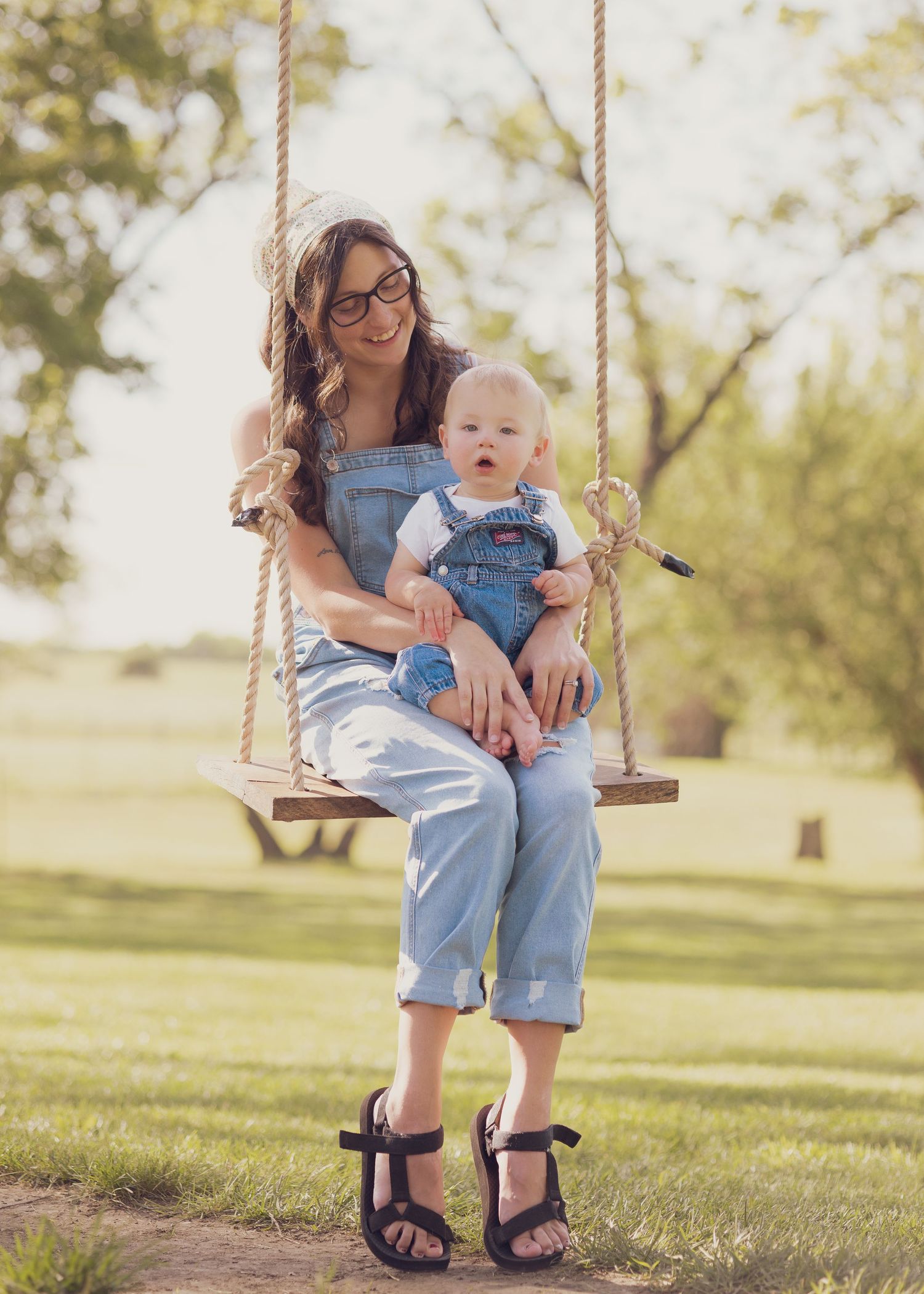 Woman in denim overalls sits on rope swing holding baby in matching overalls in sunny park.