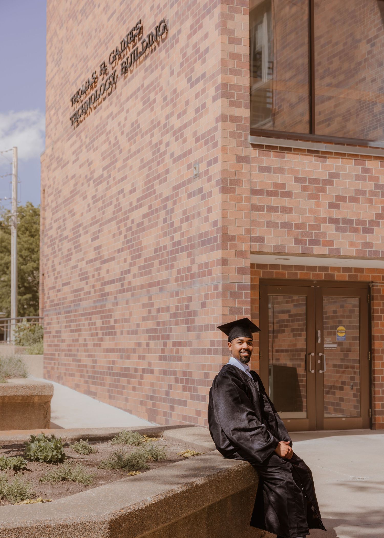 Smiling graduate in cap and gown sits outside a brick university building on graduation day.