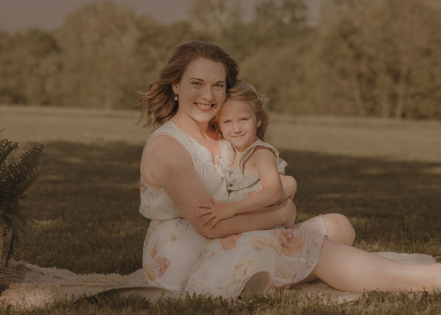 A smiling mother in a floral dress hugs her young daughter while sitting on grass in a sunlit park.