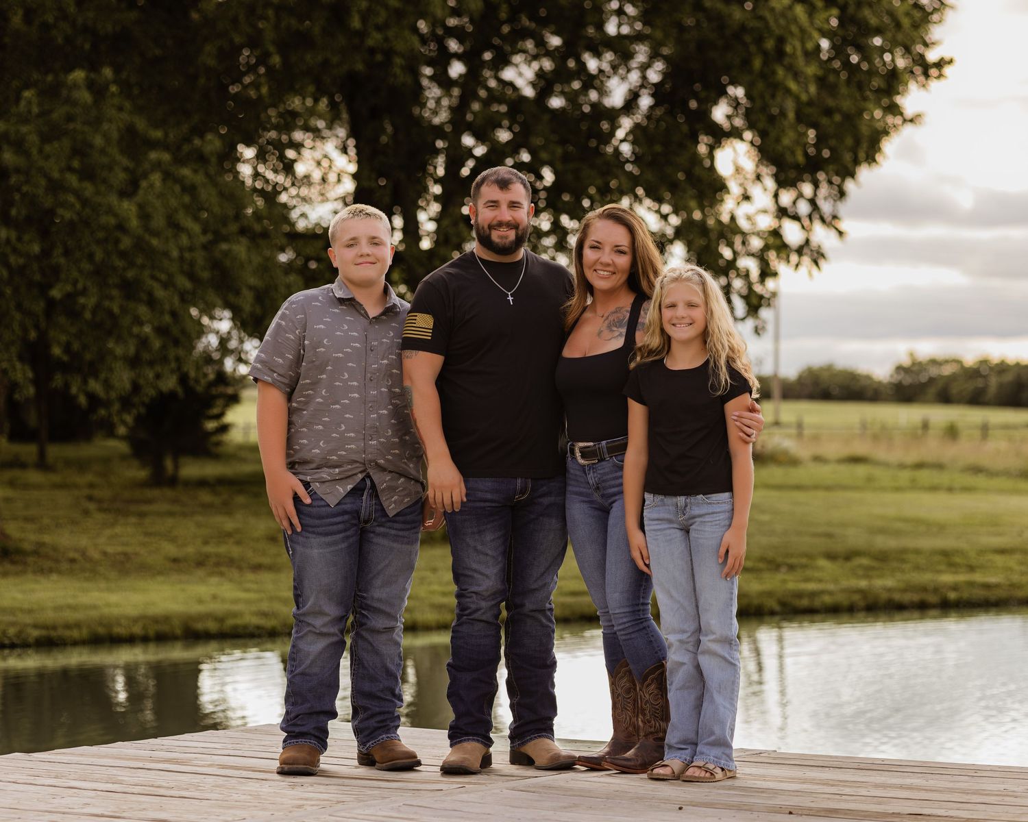 Family of four smiling together on a wooden dock beside a calm pond surrounded by green trees.