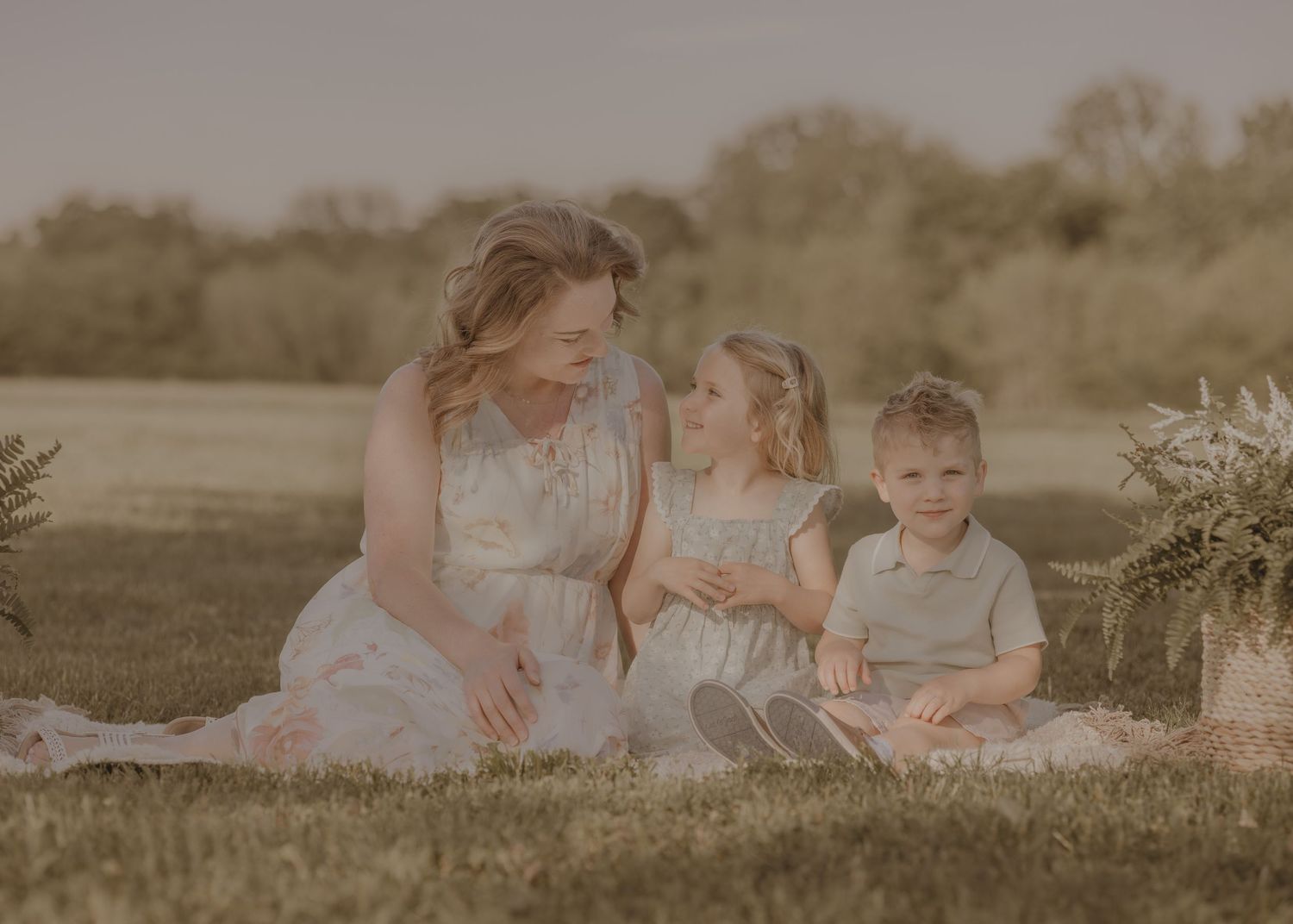 Mother sitting on grass with young daughter and toddler son during golden hour outdoor family photo session.