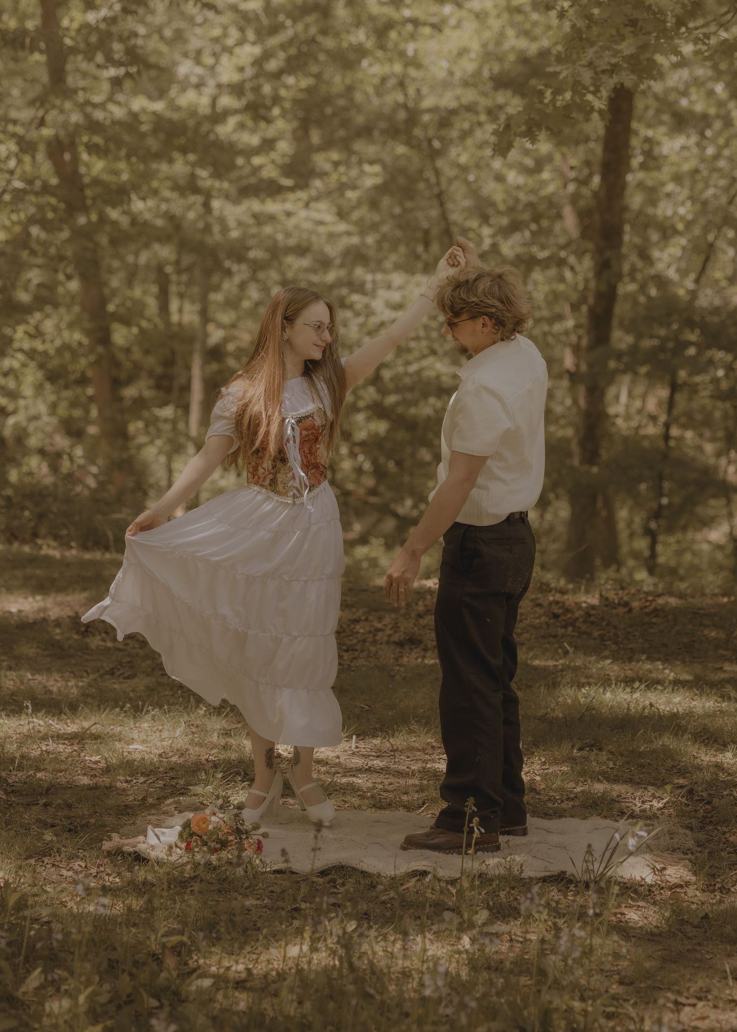 A couple dances together in a sunlit forest clearing, the woman in a white dress and the man in black pants.