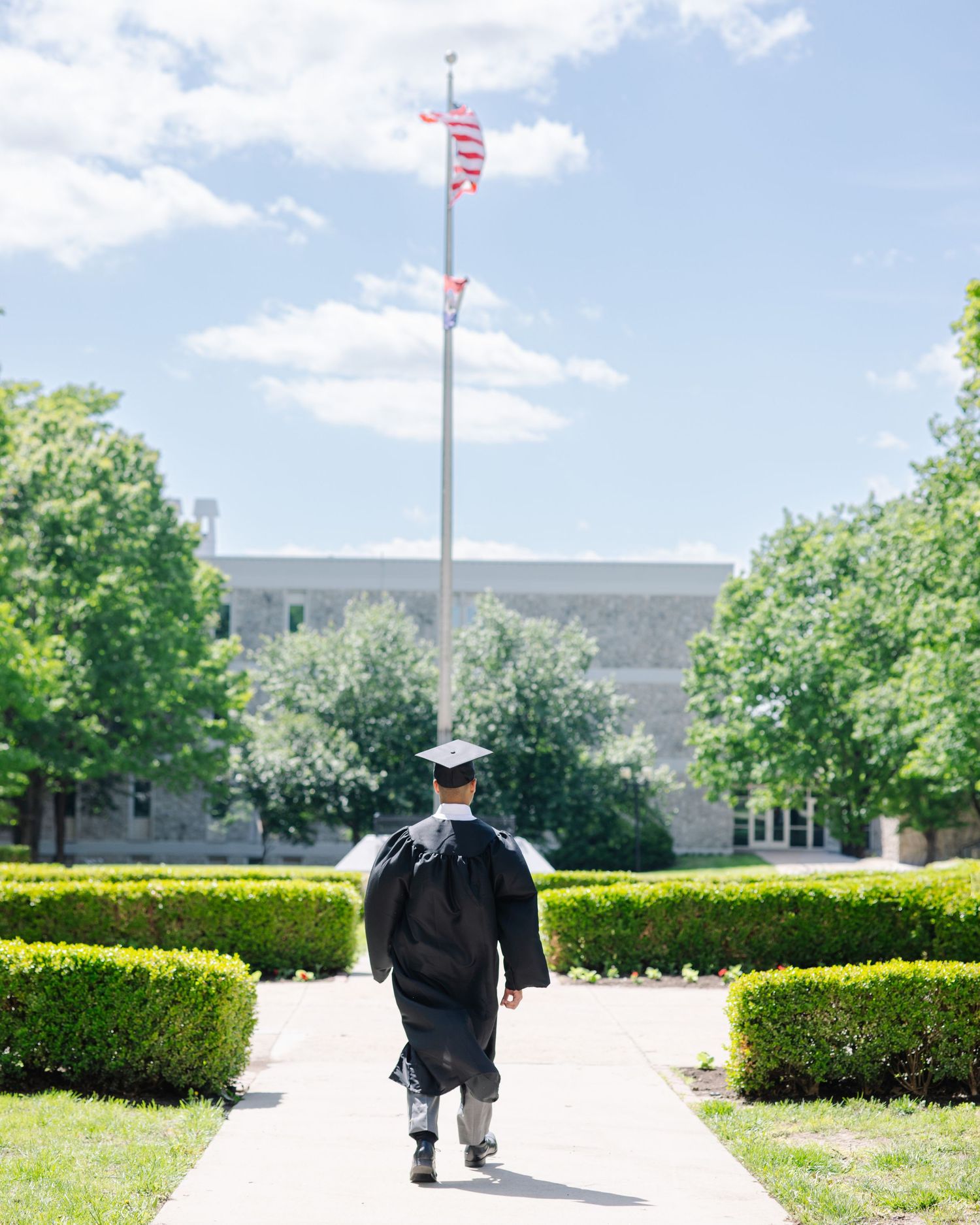 Graduate in cap and gown walks toward US flag on sunny campus pathway lined with green hedges.