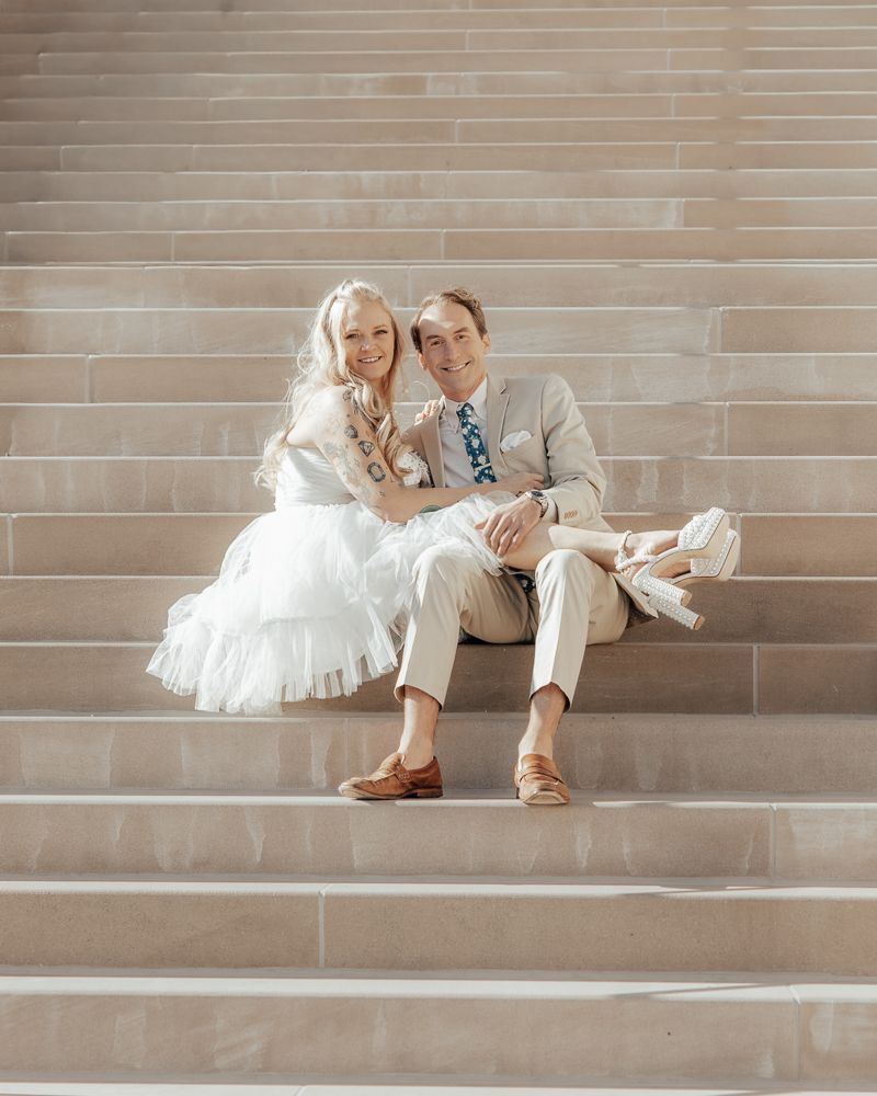 A couple sits together on stone steps, with the bride wearing a white dress and groom in light khaki attire.