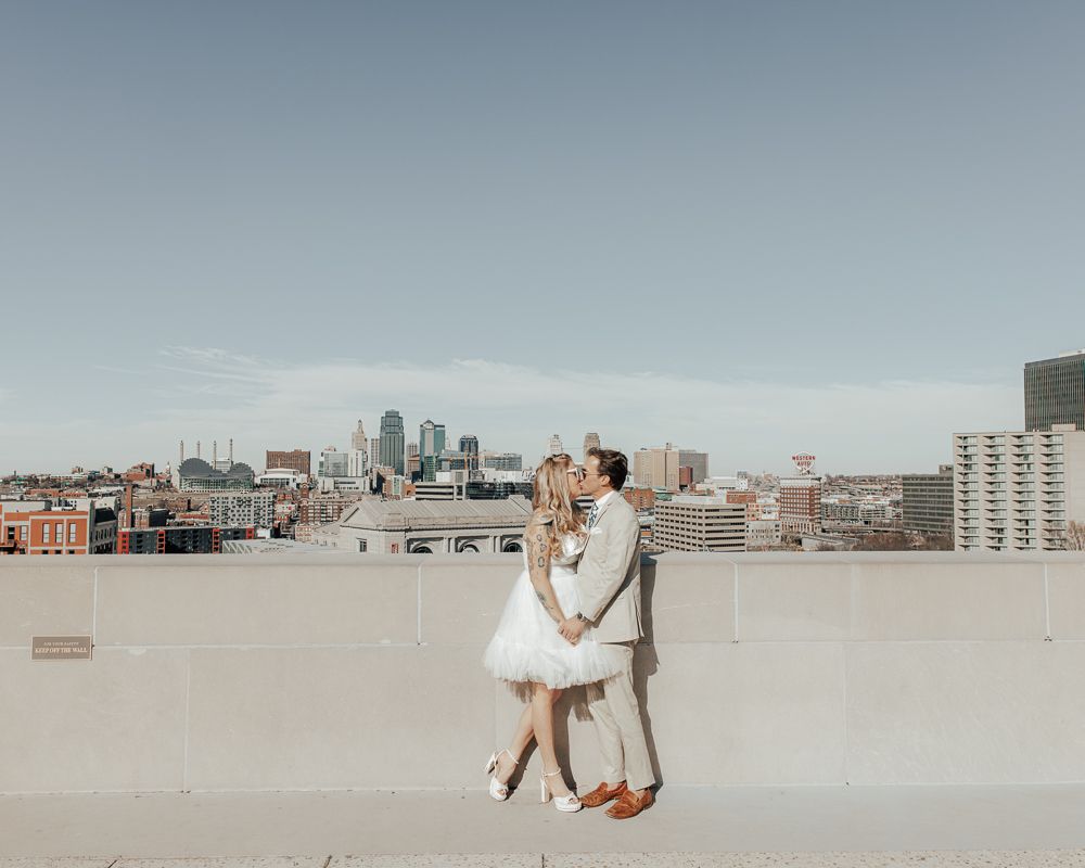 Couple in white outfits embracing on rooftop with city skyline in background.