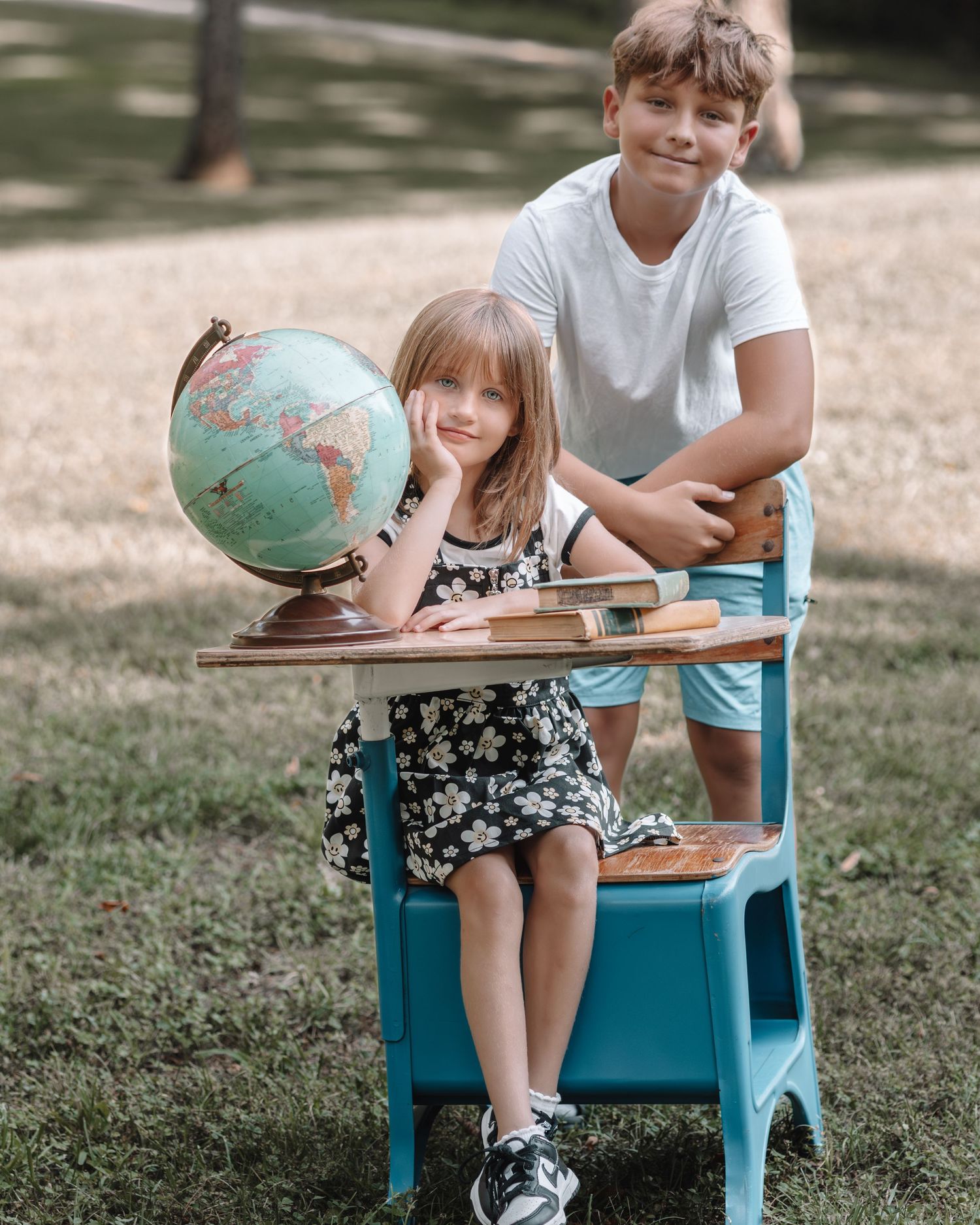 Two children pose outdoors with a globe and books on a teal school desk, capturing a back-to-school theme.