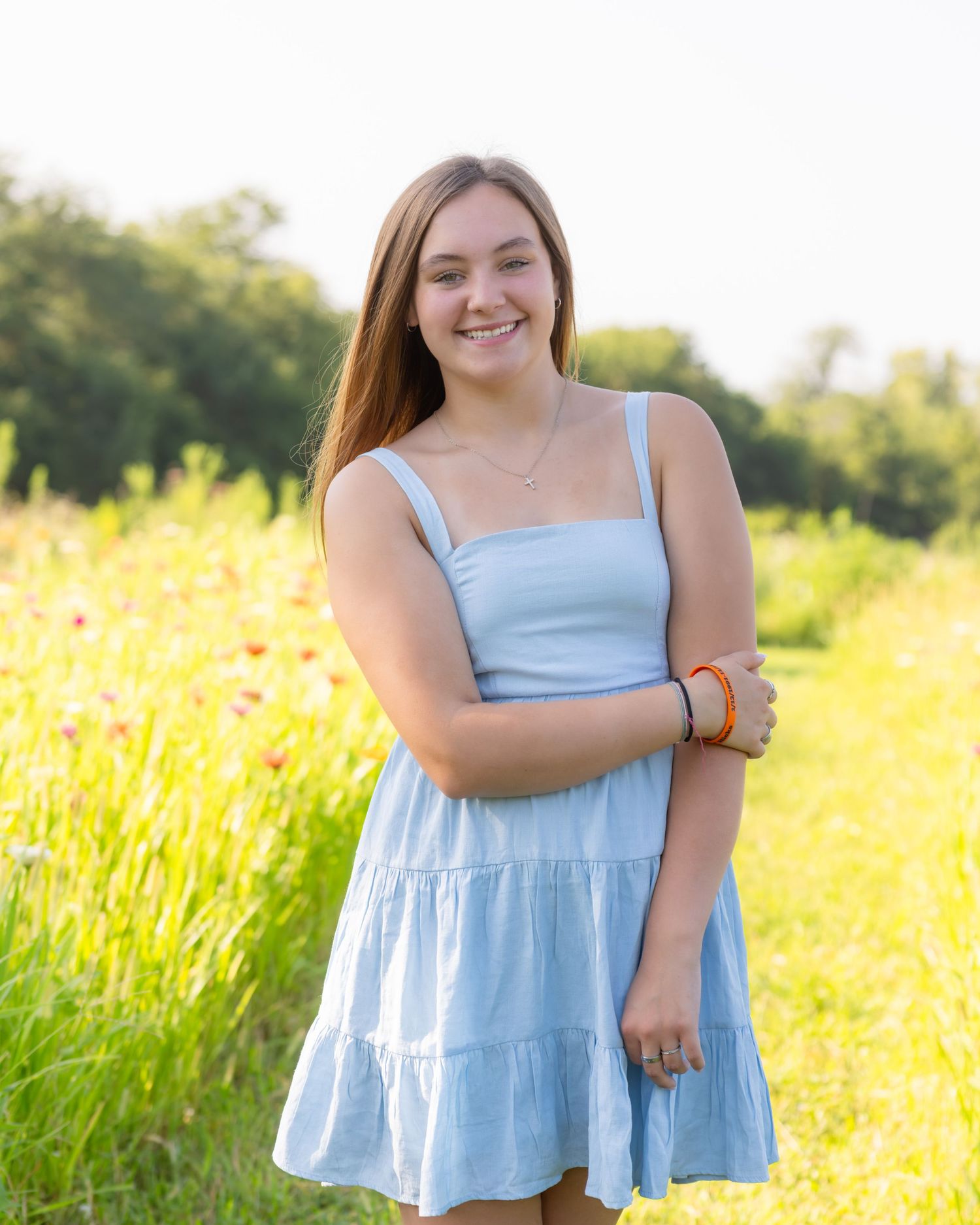 Young woman in light blue sundress smiling in a sunny wildflower field with green trees in the background.