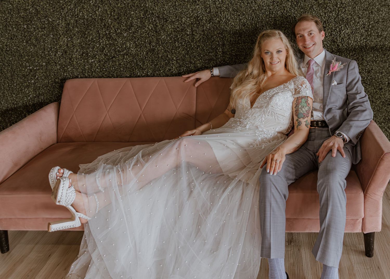Bride in white gown and groom in gray suit pose together on a pink velvet sofa at their wedding reception.