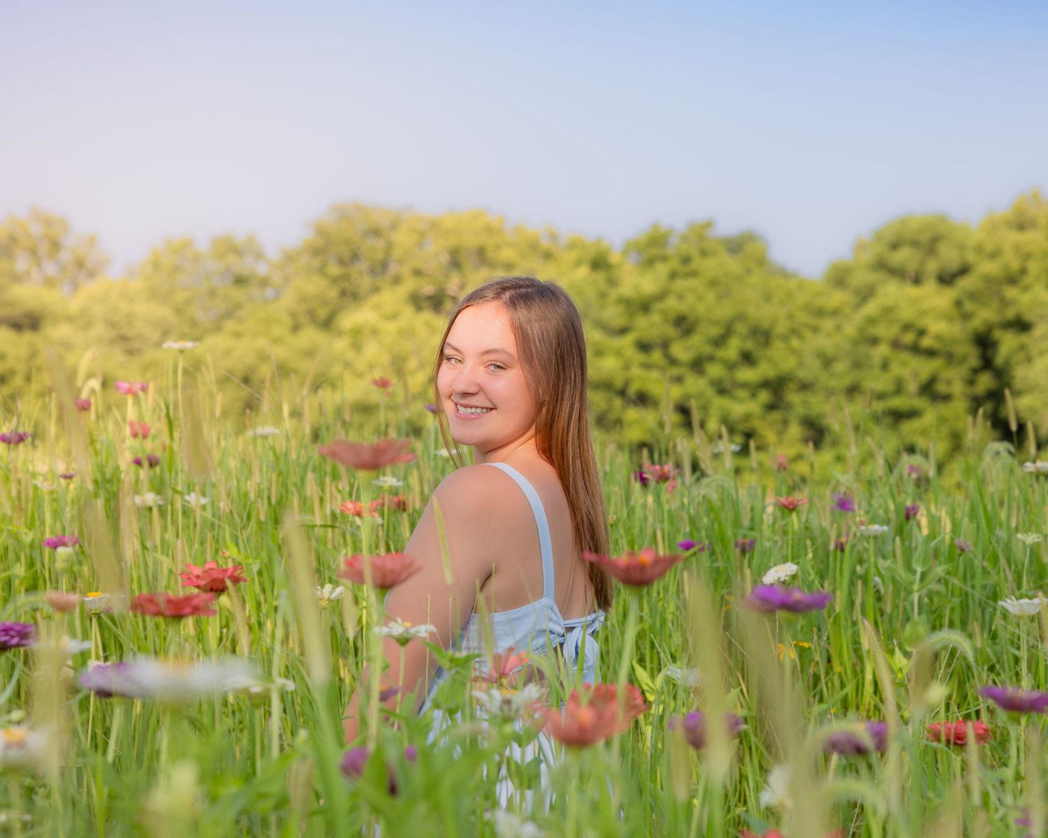 Smiling teenage girl sitting in a colorful wildflower meadow wearing a light blue dress on a sunny day.