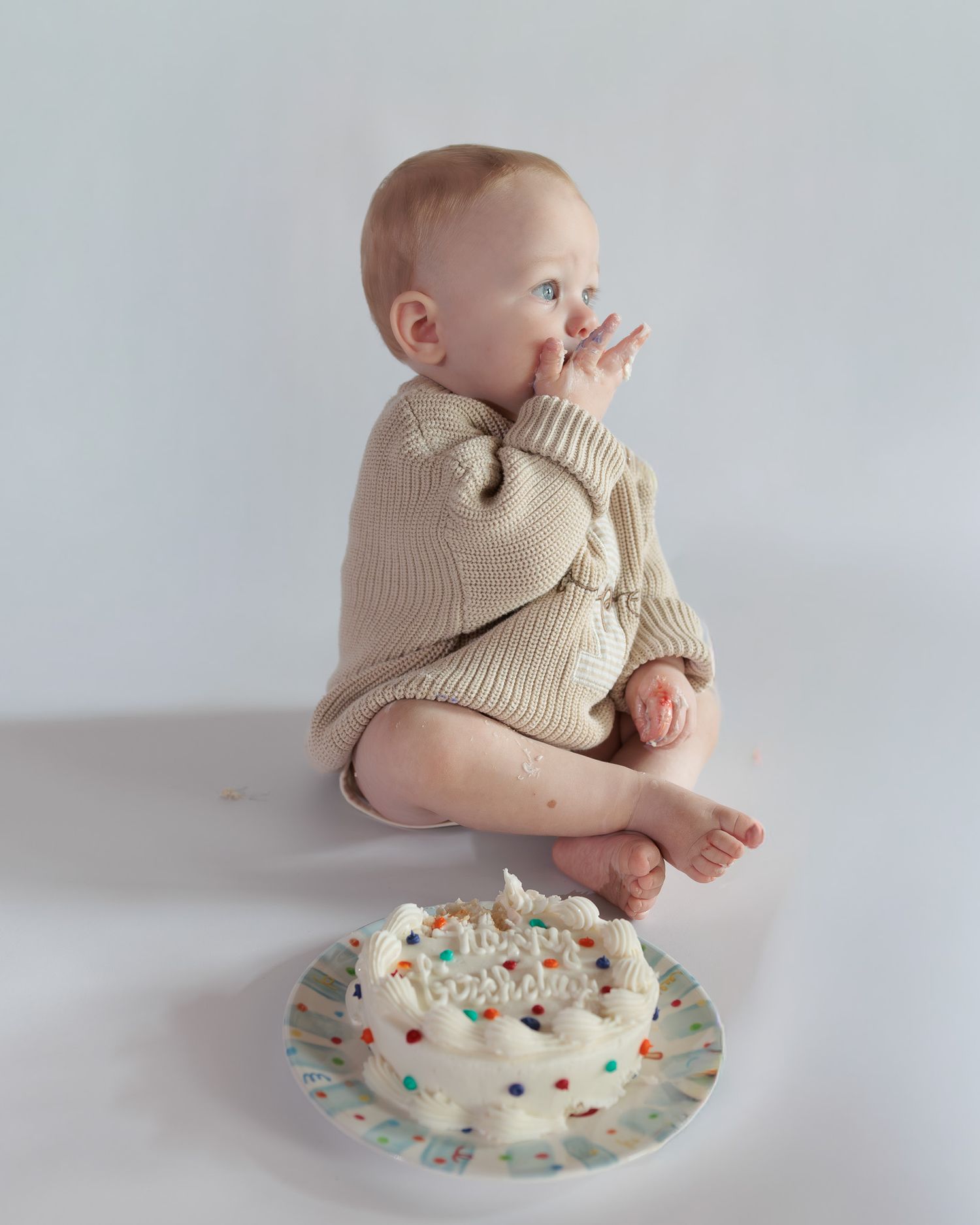 Baby in beige knit romper tasting frosting beside a white smash cake with colorful sprinkles on white background.