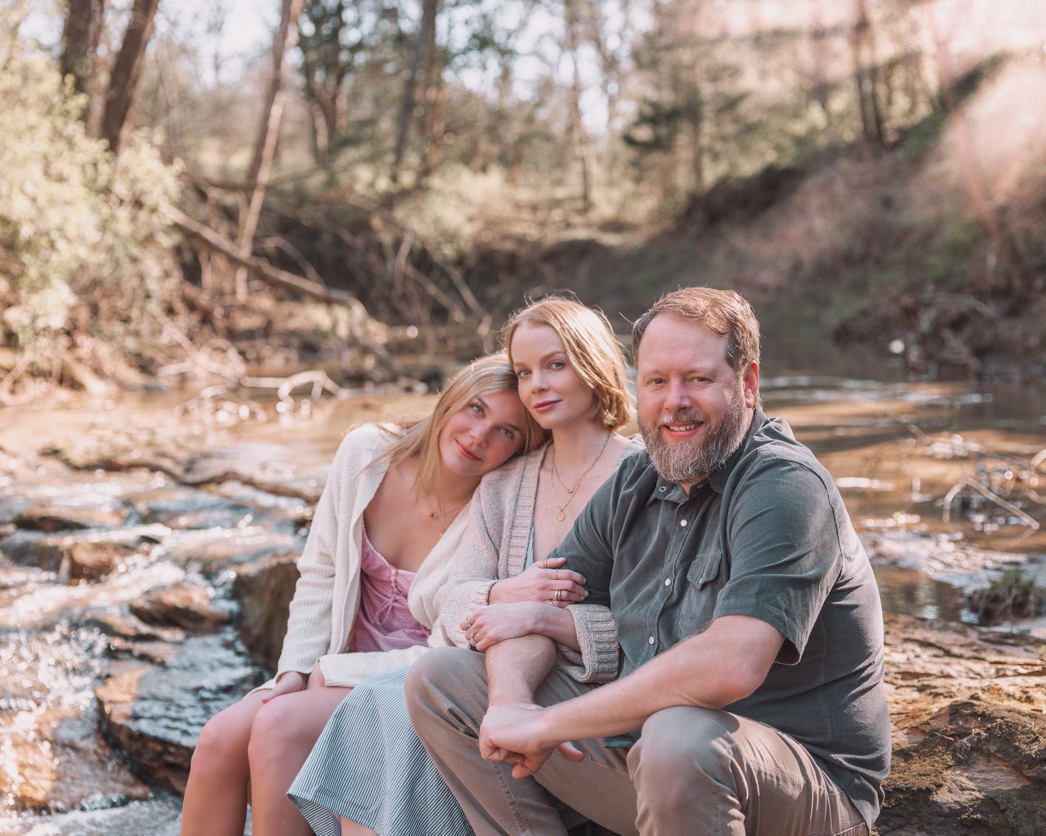 Family of three posing by a rocky creek in a sunlit forest, smiling warmly for an outdoor portrait photo.