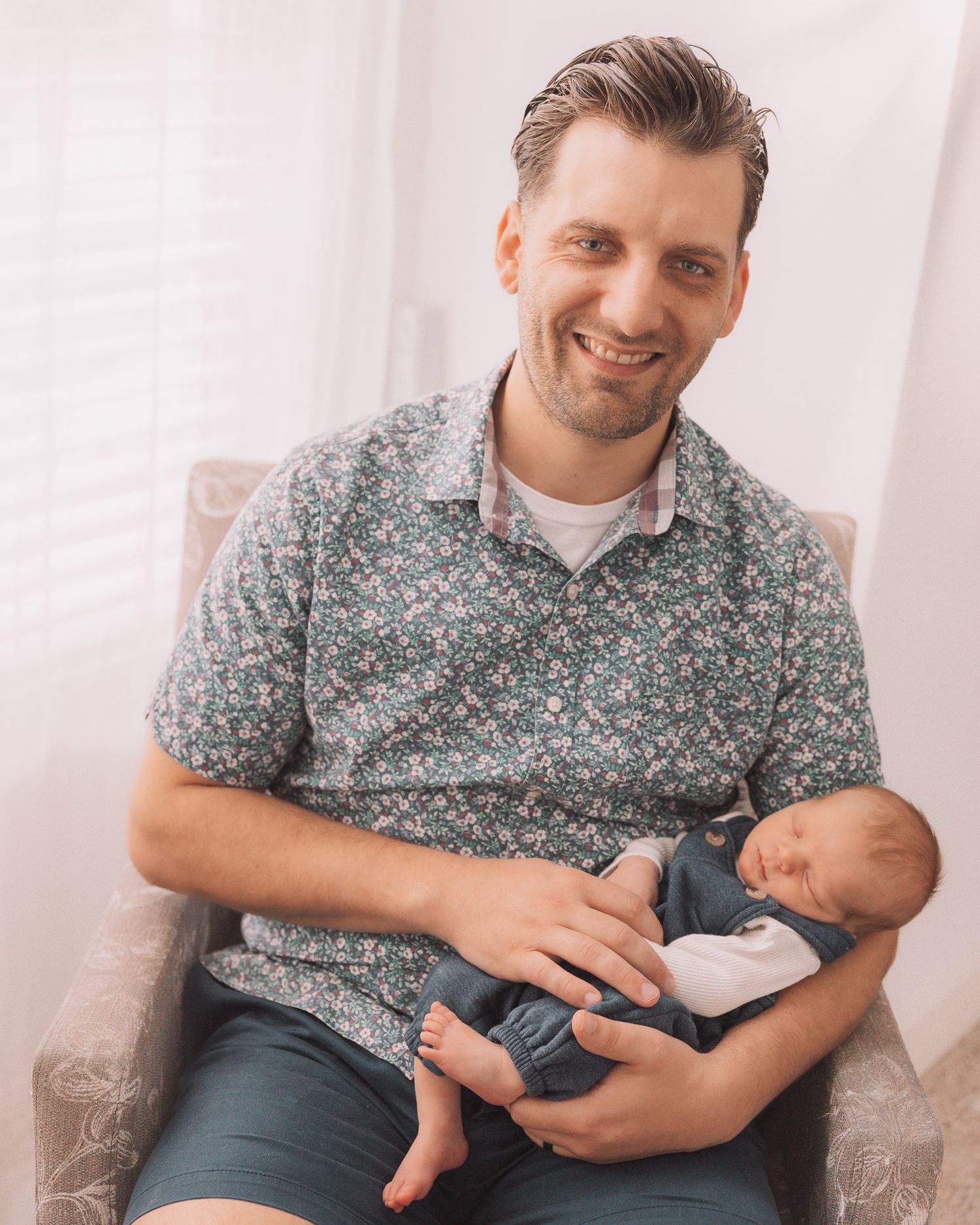 Smiling man in a floral shirt sitting in a chair, gently cradling a sleeping newborn baby.