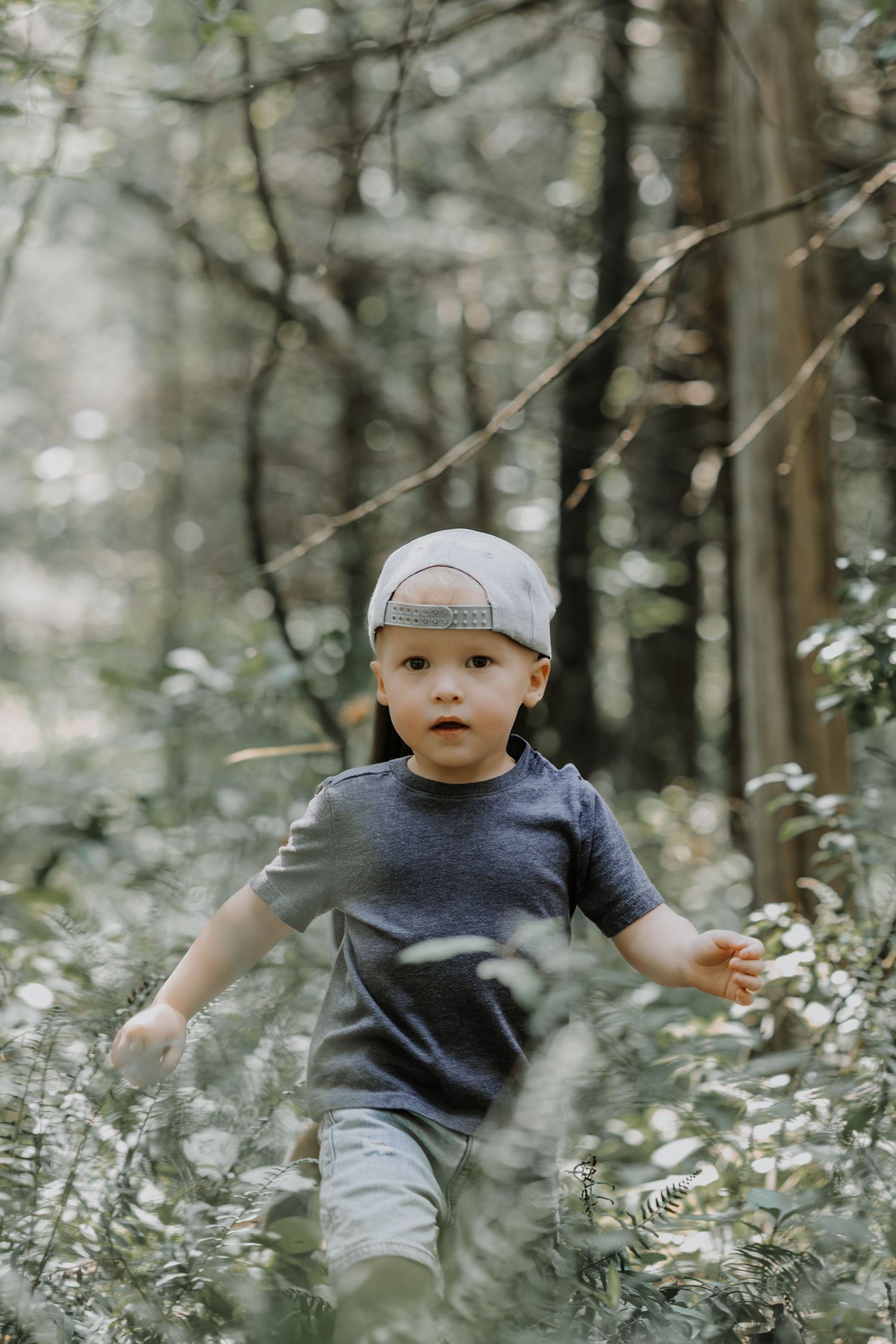 A small child wearing a baseball cap and dark t-shirt explores through a misty forest with white wildflowers.
