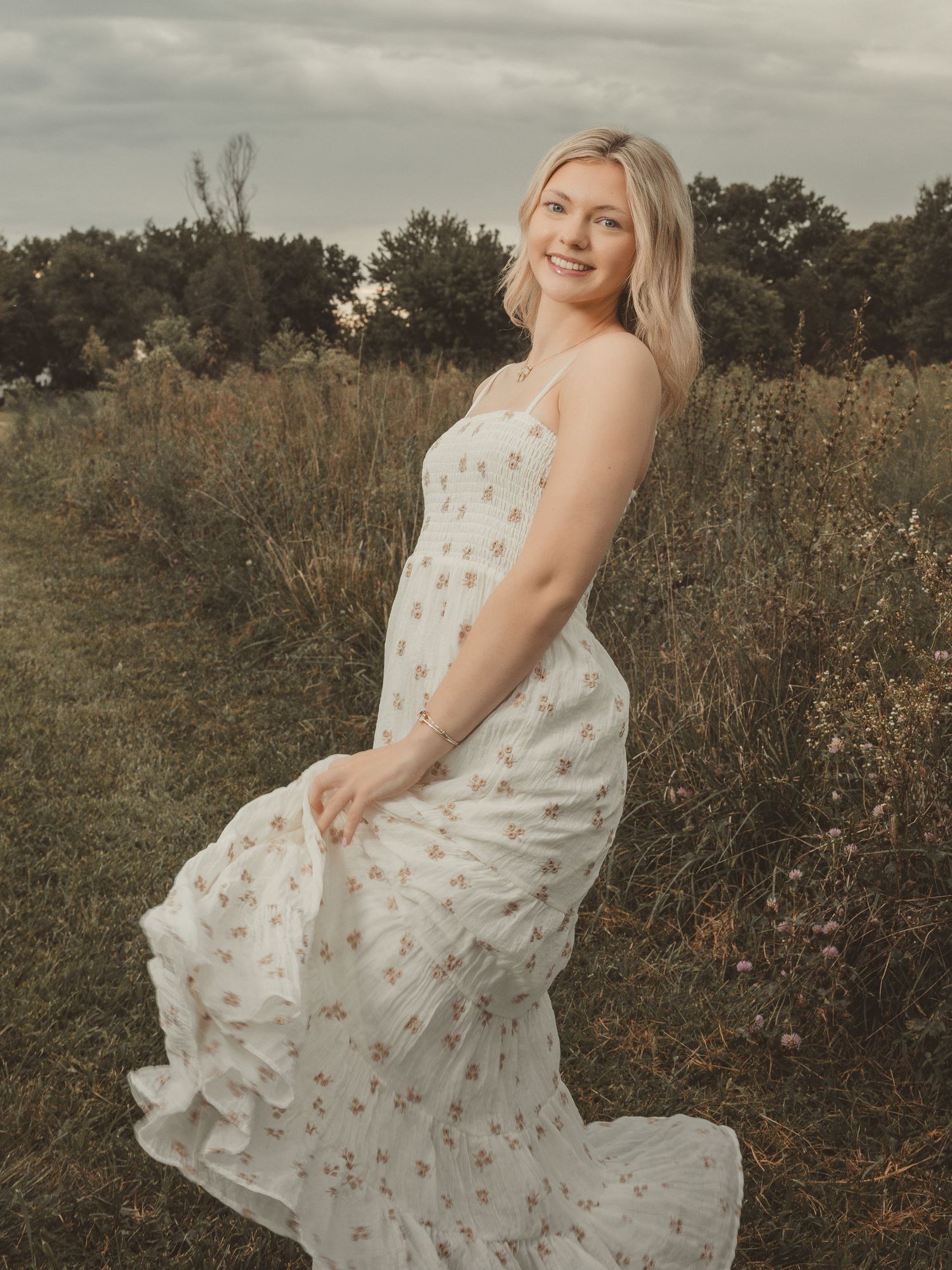 Blonde woman in floral white maxi dress posing gracefully in a meadow field on an overcast day.
