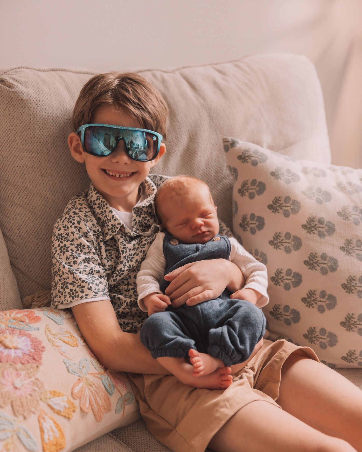 A young boy wearing blue sunglasses holds a sleeping newborn baby while sitting on a couch.