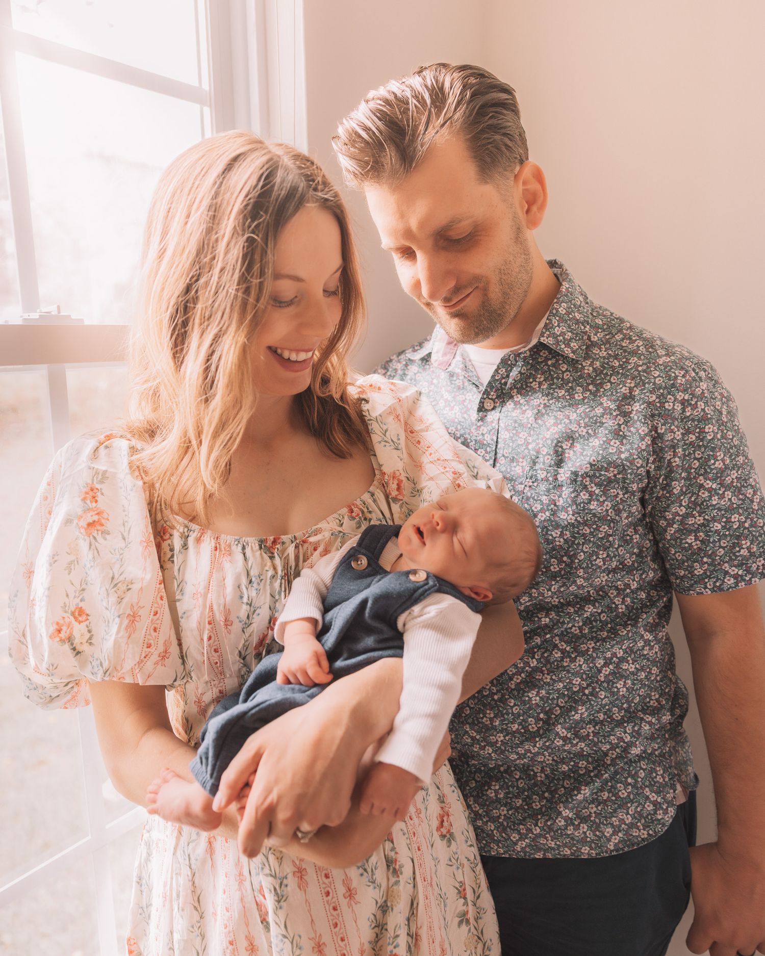 Happy couple smiling and holding their newborn baby by a bright window, wearing floral outfits in a warm, soft-lit room.