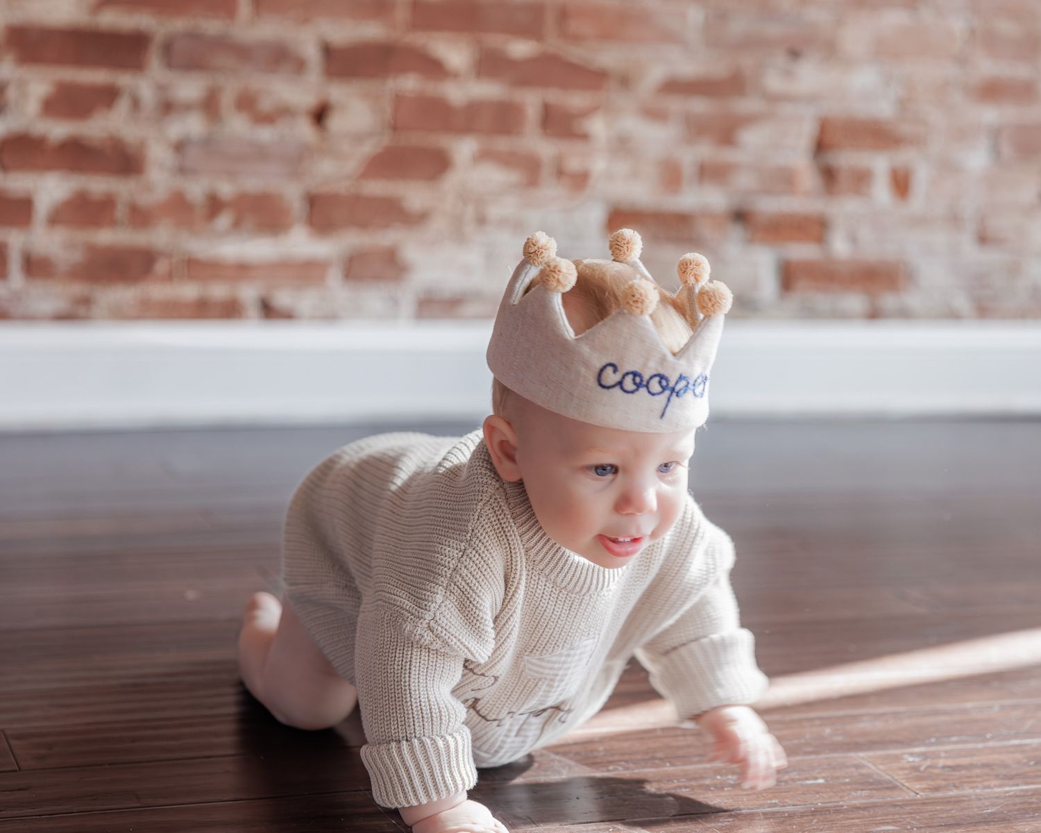 Baby wearing personalized name crown crawling on hardwood floor in cream knit romper against brick wall.