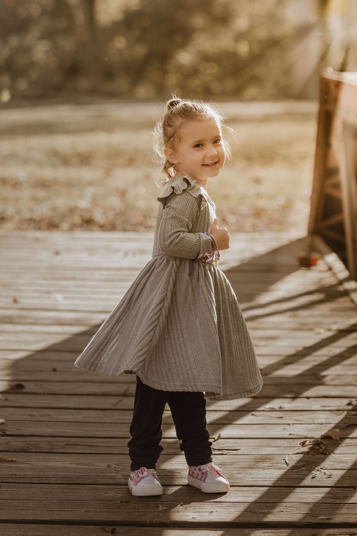 A small child in a gray dress twirls and plays on a wooden deck during golden hour, casting long shadows.