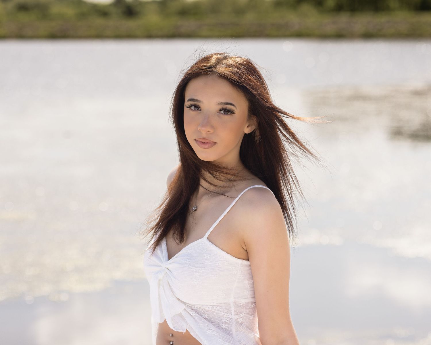 Young woman with brown hair in white crop top posing near a serene lake on a bright sunny day.
