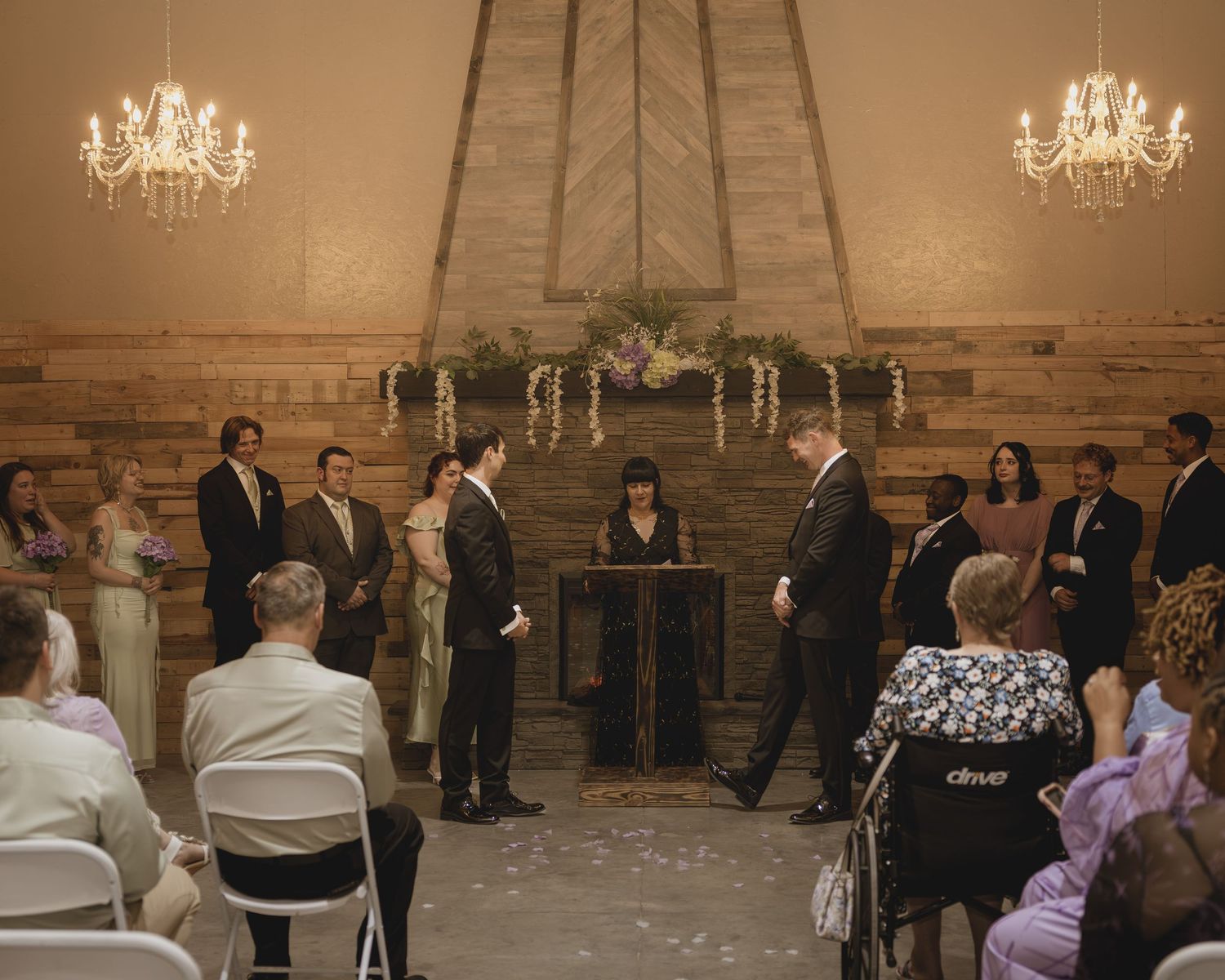 Wedding ceremony indoors with couple at altar, officiant, wedding party, and seated guests under chandelier lighting.