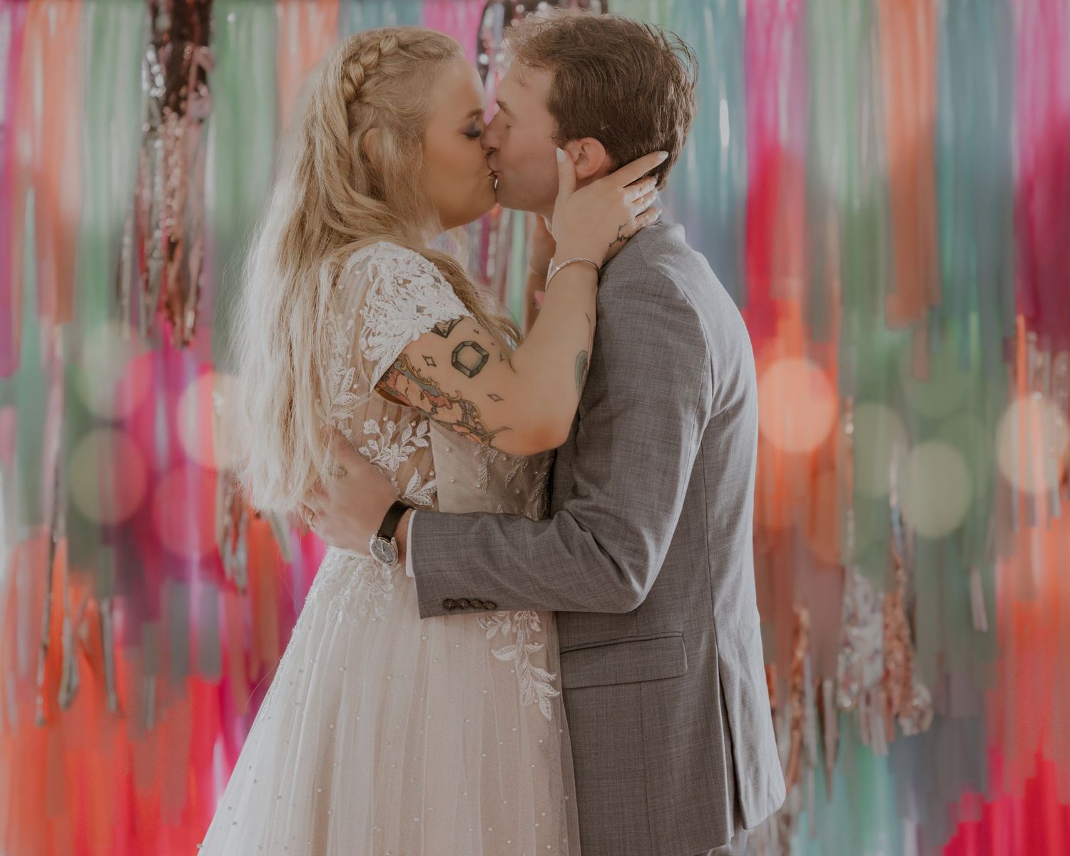 Bride and groom share first kiss at wedding altar with colorful painted backdrop in soft bokeh lighting.