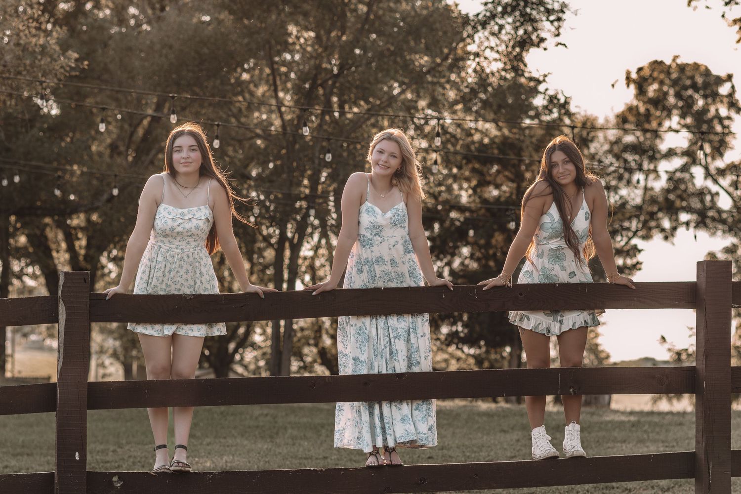 Three young women in floral dresses pose on a rustic wooden fence at golden hour in a scenic outdoor setting.