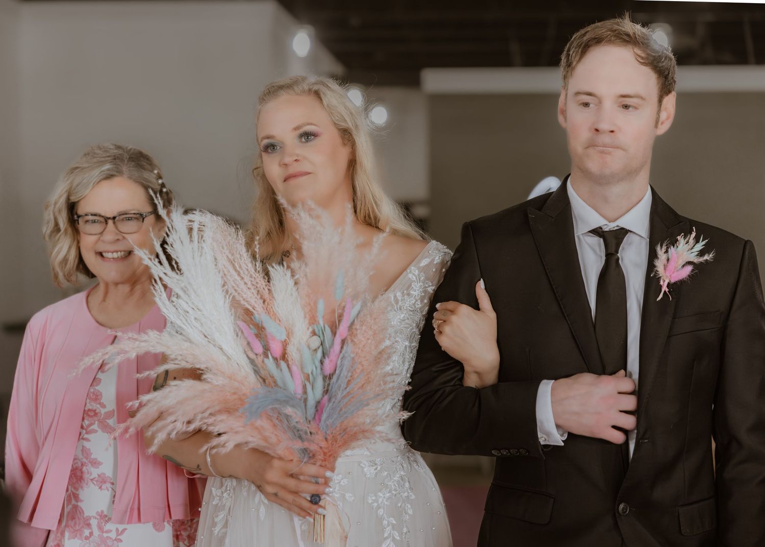 Bride in white gown holding colorful feather bouquet with groom in suit and older woman in pink at wedding.