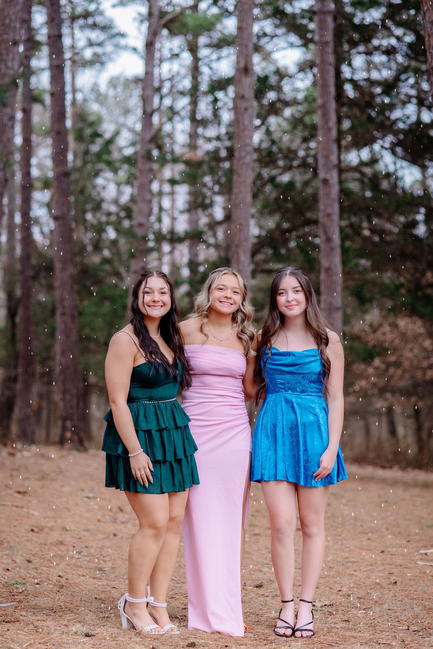Three friends in colorful formal dresses pose together in a wooded setting on a dirt path.