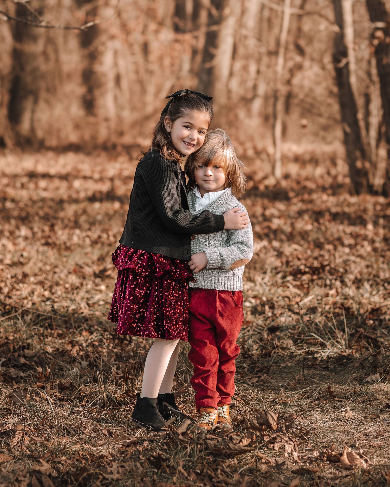 Two young children in coordinated outfits share a sweet moment together in a fall forest setting.