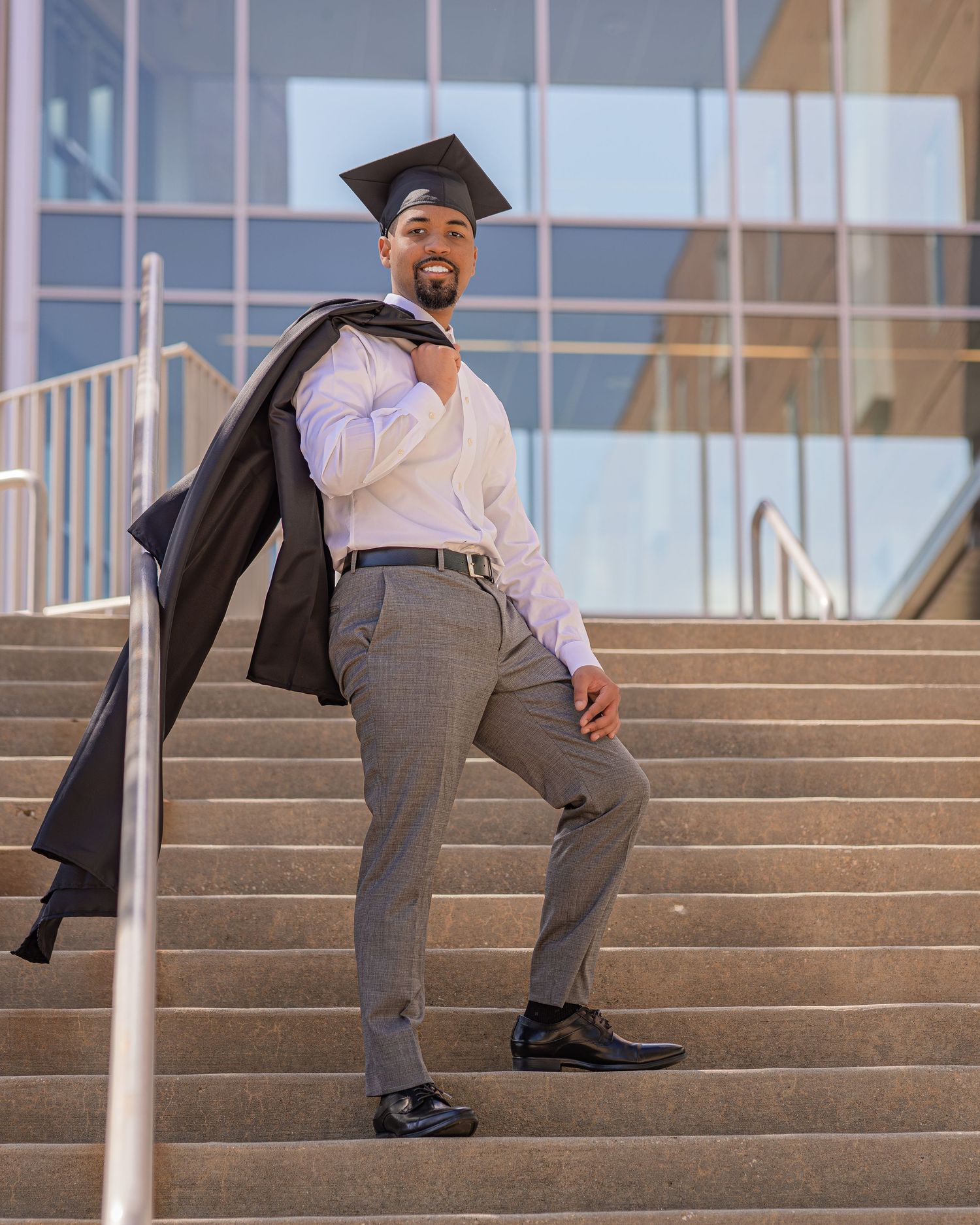 Graduate smiling on steps holding gown over shoulder wearing cap and dress clothes.