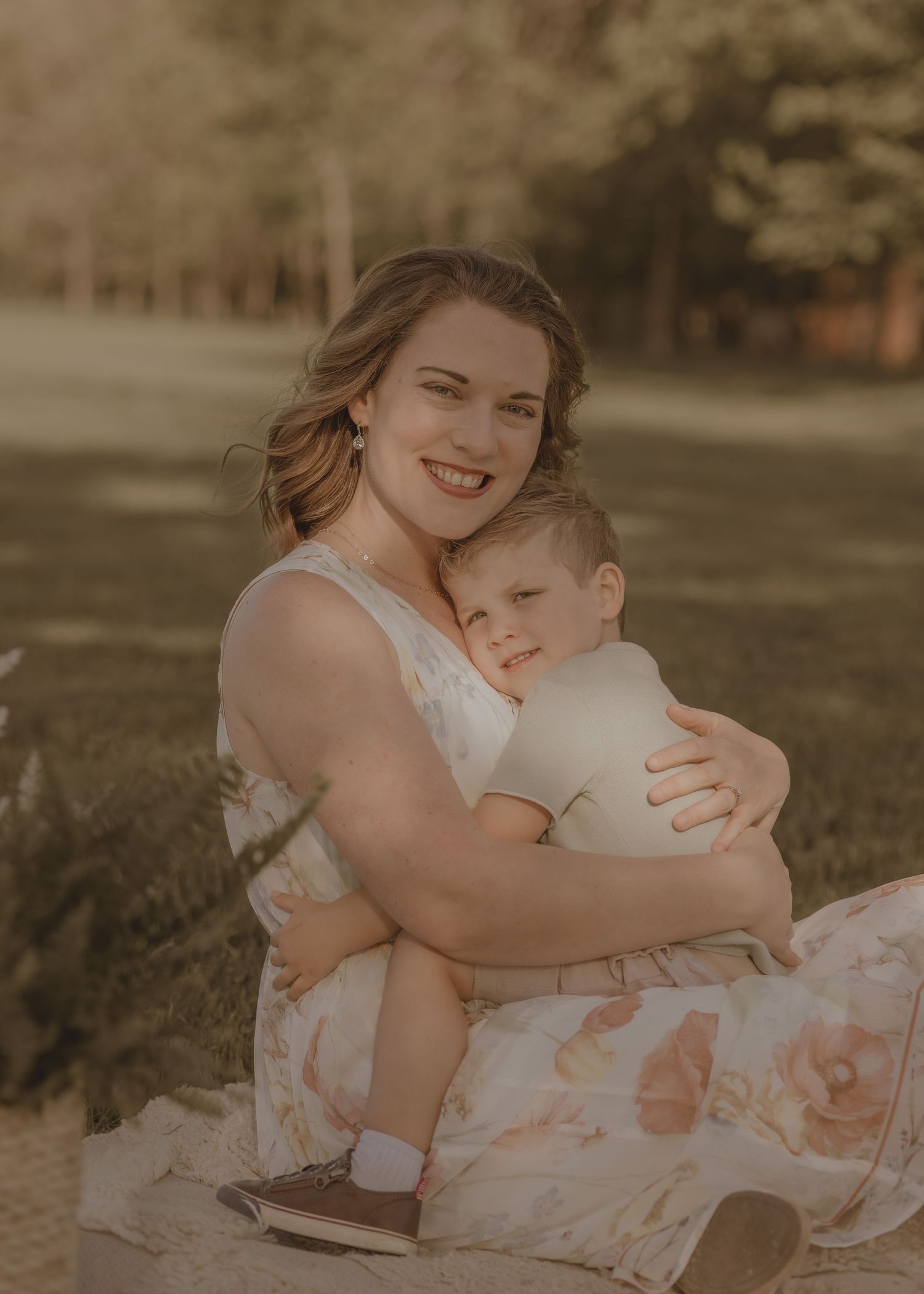 A smiling mother in a floral dress holds her baby outdoors in a warm, golden-lit park setting.