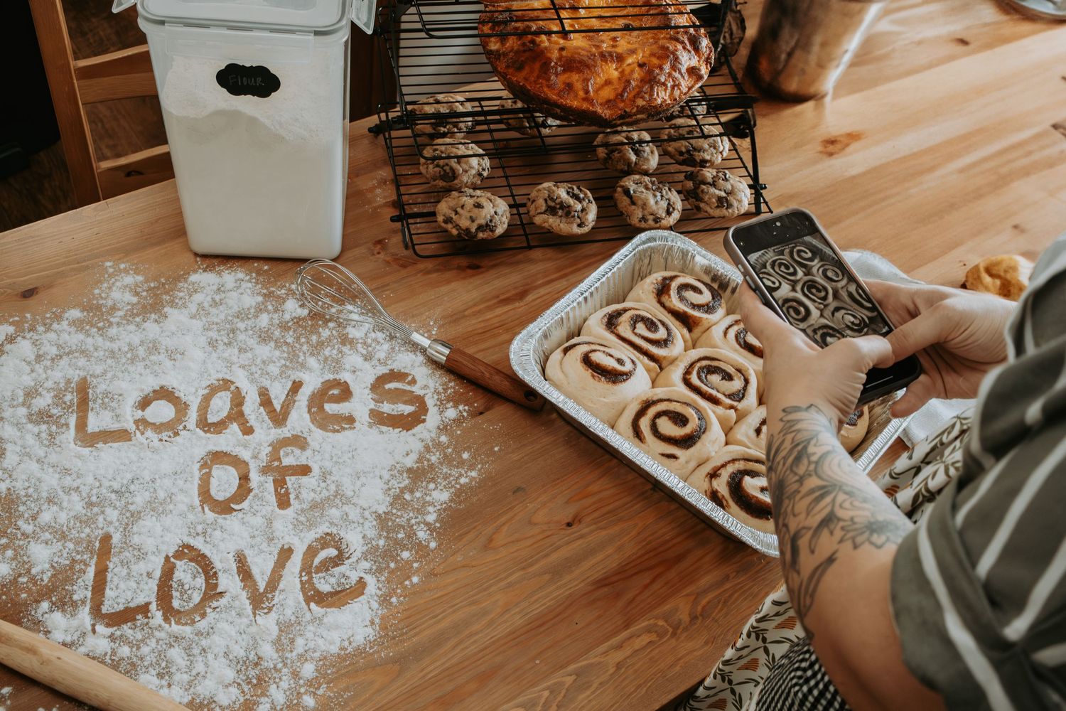 Baker photographing fresh cinnamon rolls on a tray beside flour spelling Loaves of Love on a wooden surface.
