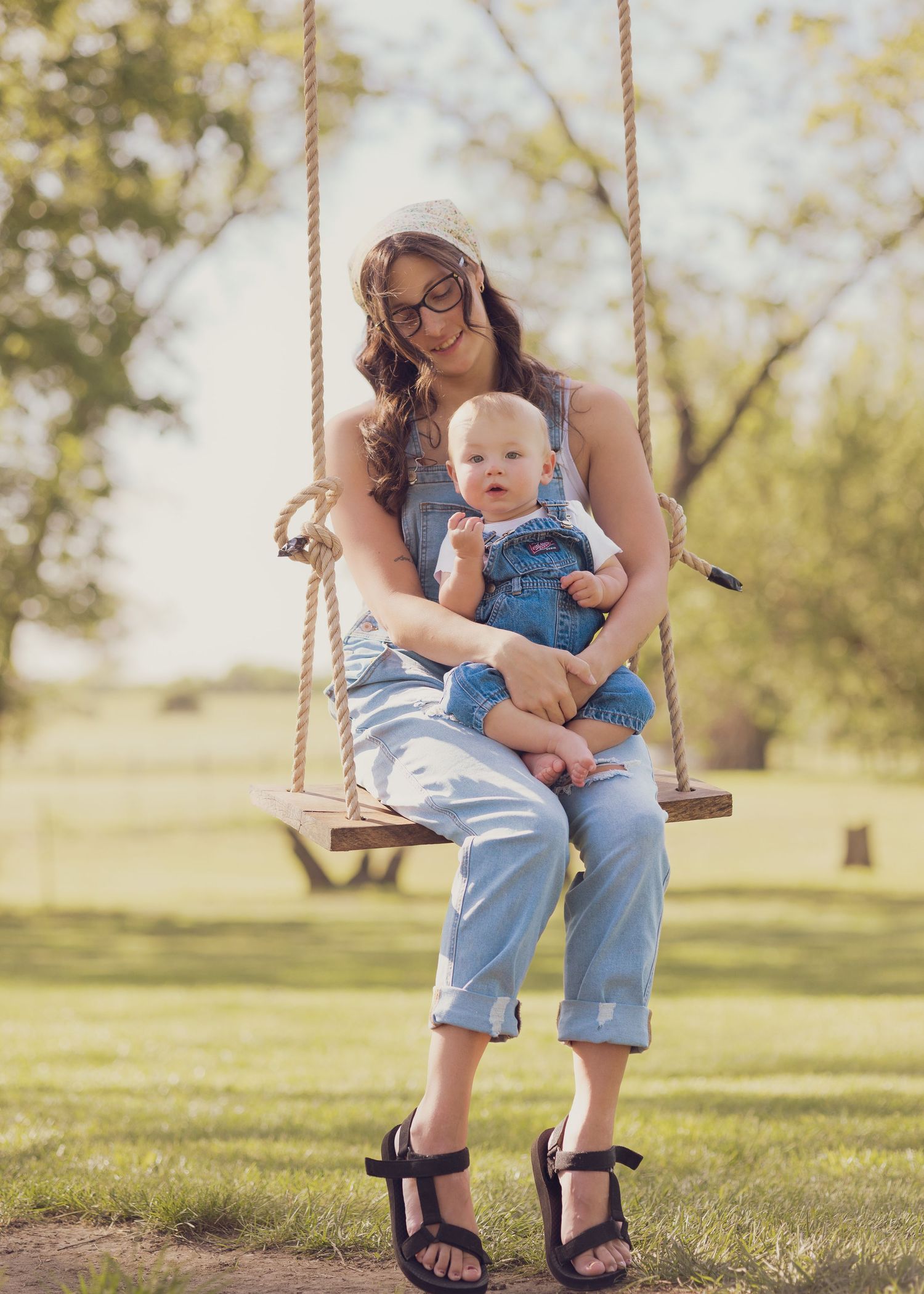 A young mother in denim overalls sits on a rope swing holding her baby dressed in matching denim overalls outdoors.