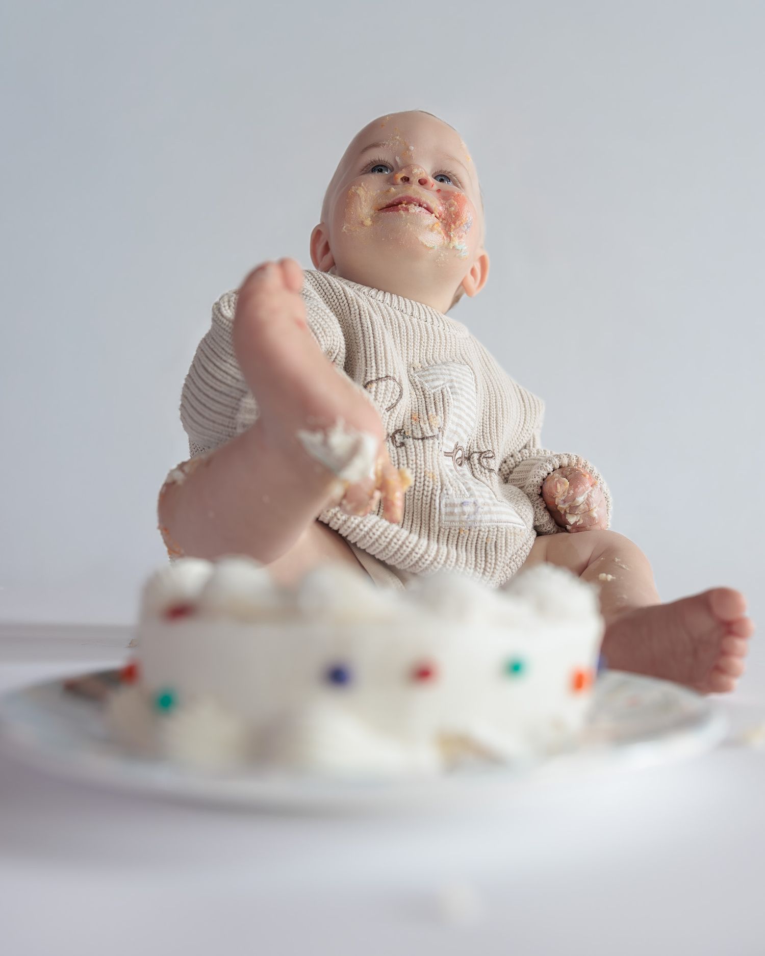 Baby covered in frosting sits behind smashed first birthday cake with colorful sprinkles on white background.