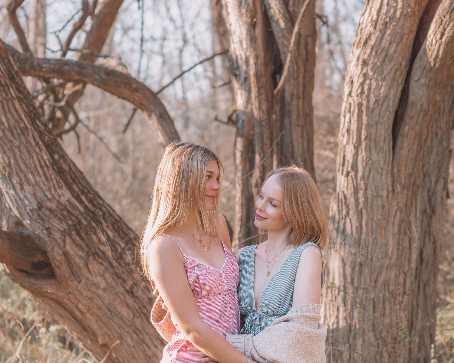 Two young women in pastel dresses share a tender moment leaning against a large tree in a natural outdoor setting.