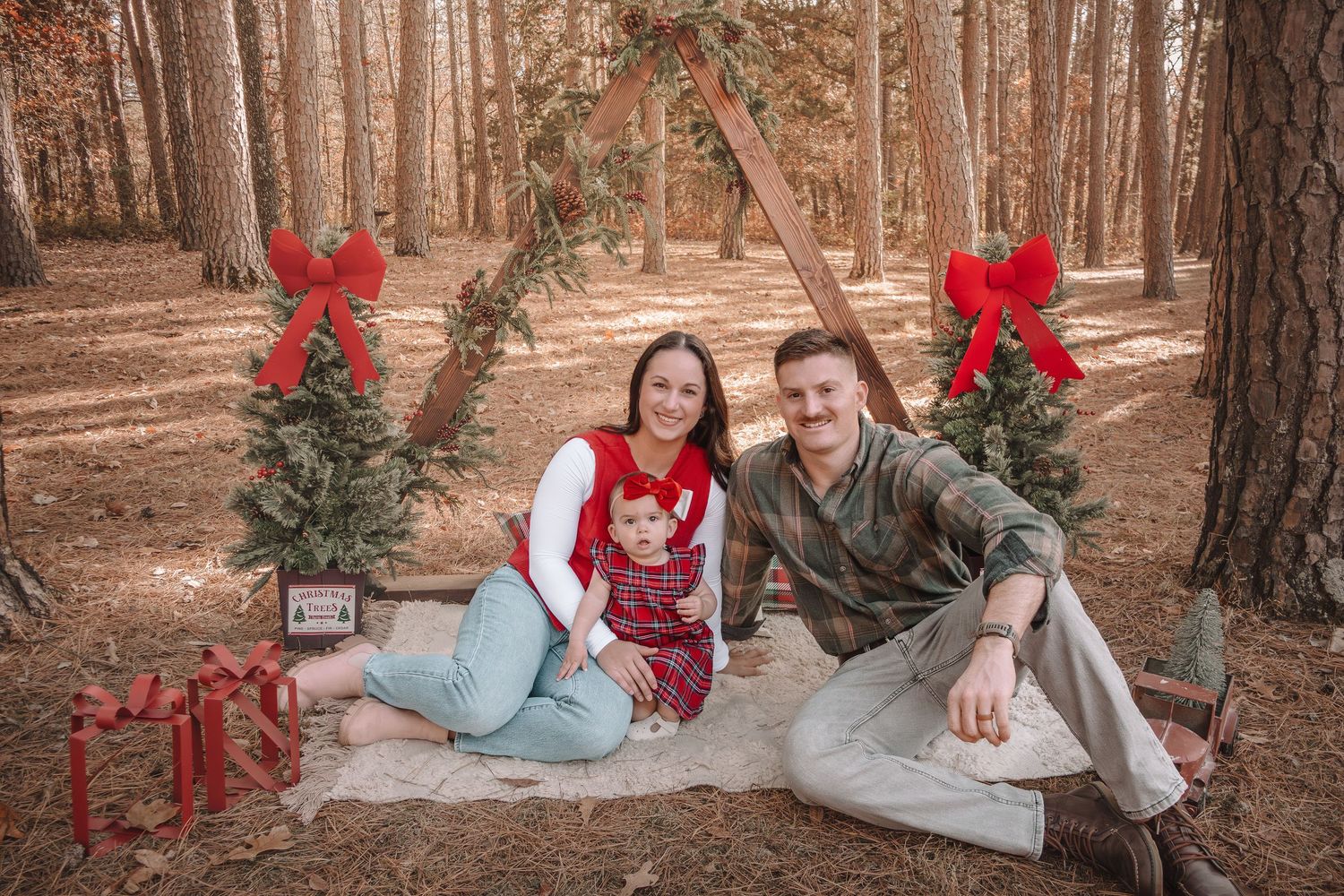 Holiday portrait in pine forest with couple sitting on blanket between two decorated Christmas trees with red bows.