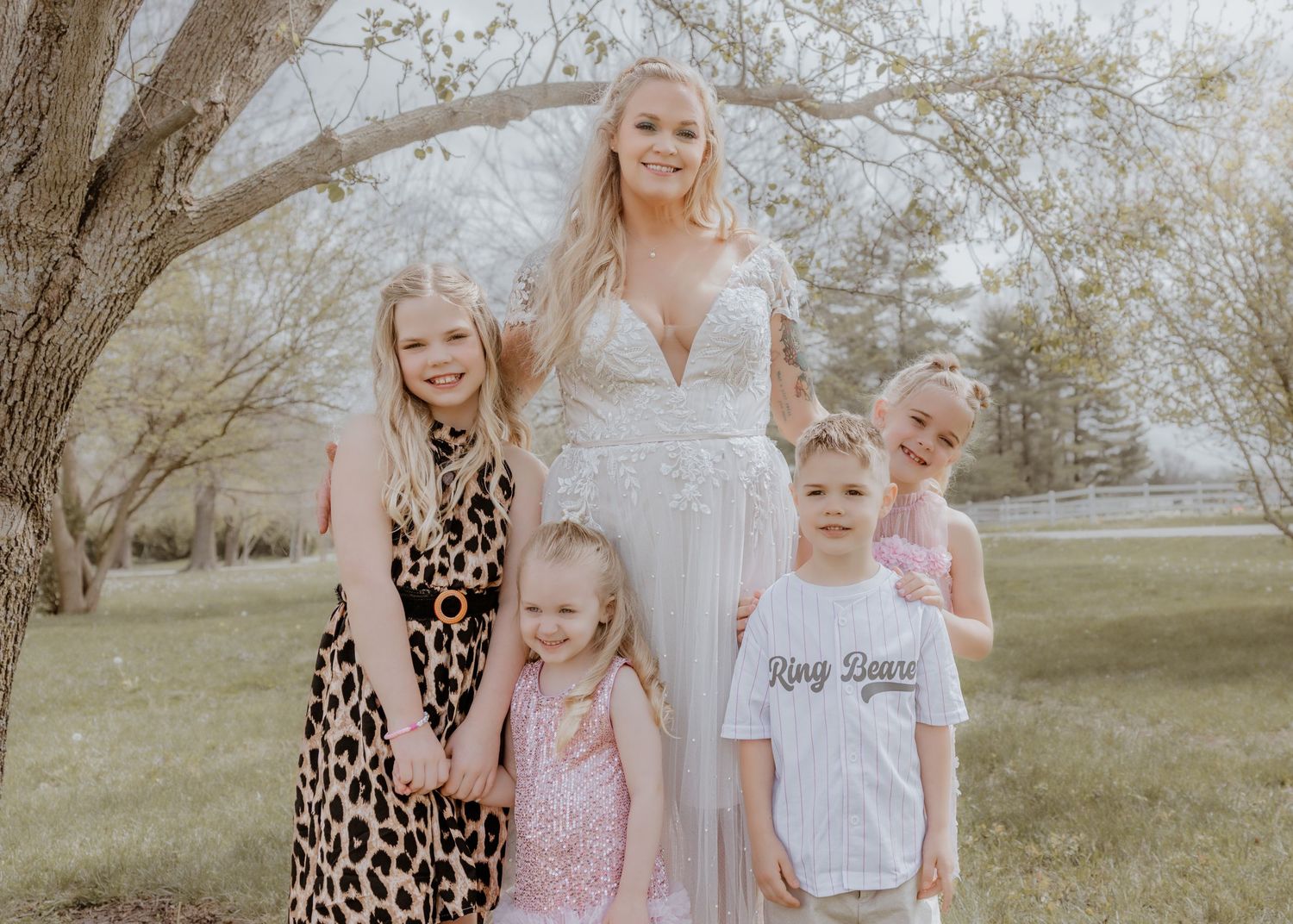 Bride in white lace gown poses outdoors with four young children under blooming spring trees.