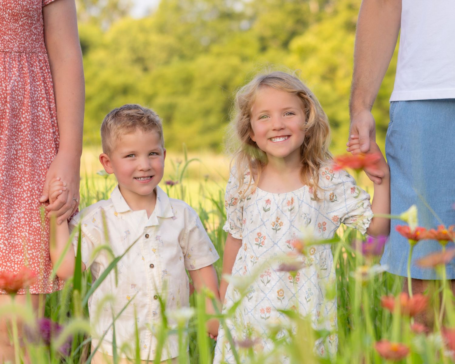Two young siblings smile in a sunny wildflower field, holding hands with parents in a bright outdoor family portrait.