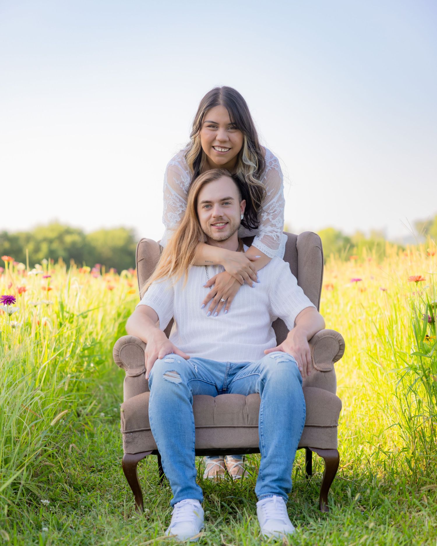 Couple posing outdoors in a sunny wildflower field, woman standing behind man seated in a wingback chair.