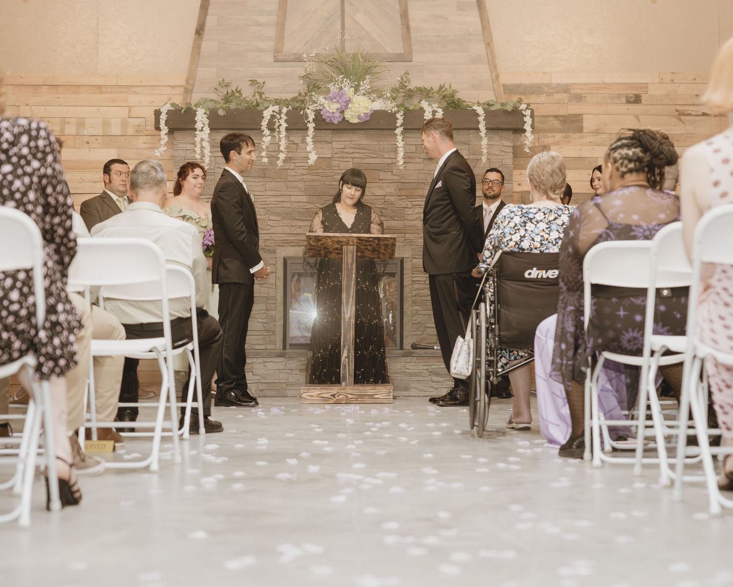 Elegant indoor wedding ceremony with couple exchanging vows at altar, guests seated in white chairs, rustic wood backdrop.