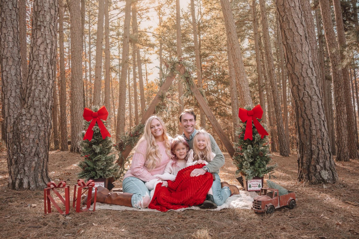 A family sits together on a red bench in a sunlit forest setting decorated with holiday bows and decor.