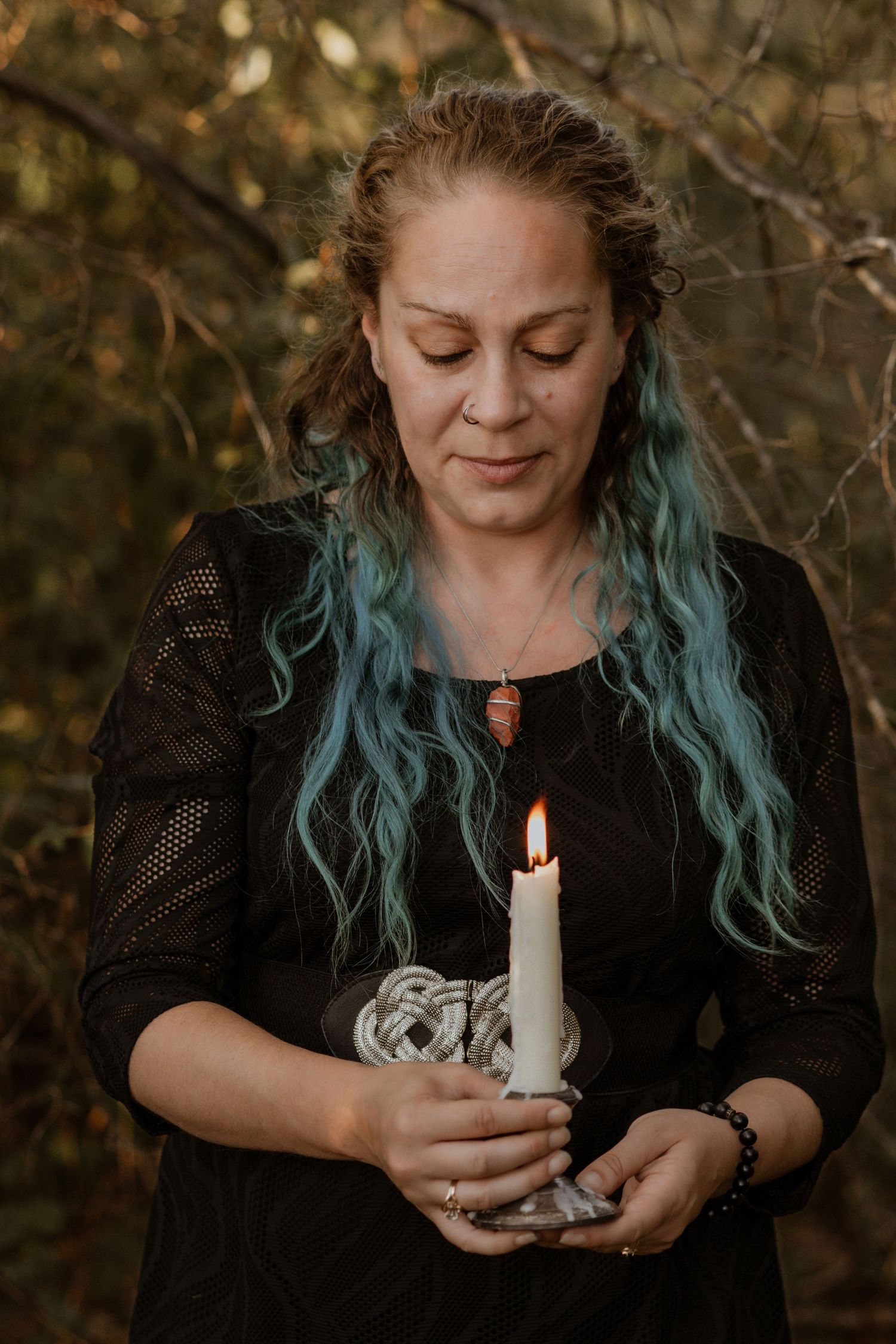 Person with blue hair holding a lit white candle in an outdoor autumn setting.