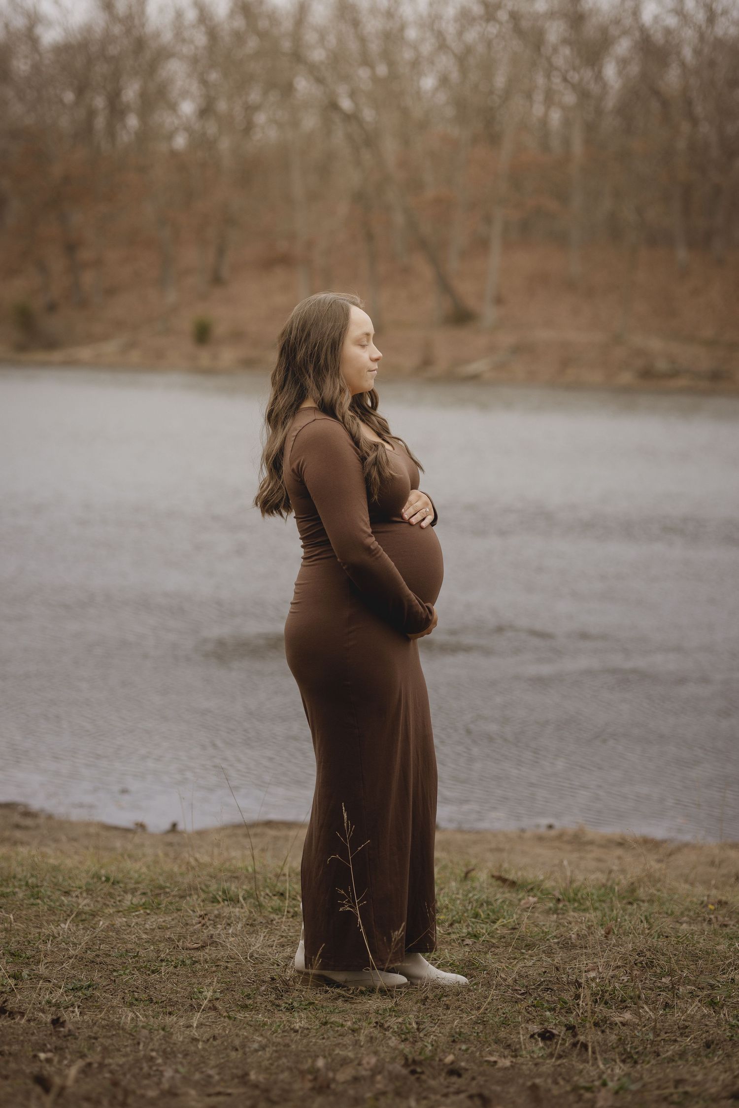 Expectant mother in a brown dress stands by a lake during a maternity photoshoot.
