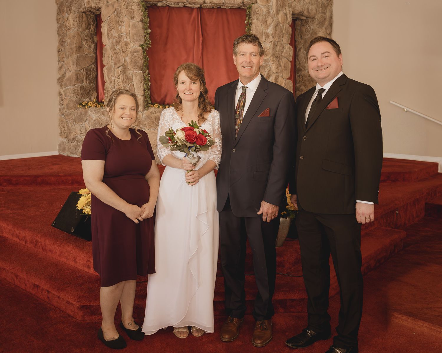A formal wedding photo of a wedding party of four people standing on red carpeted stairs in a traditional venue.
