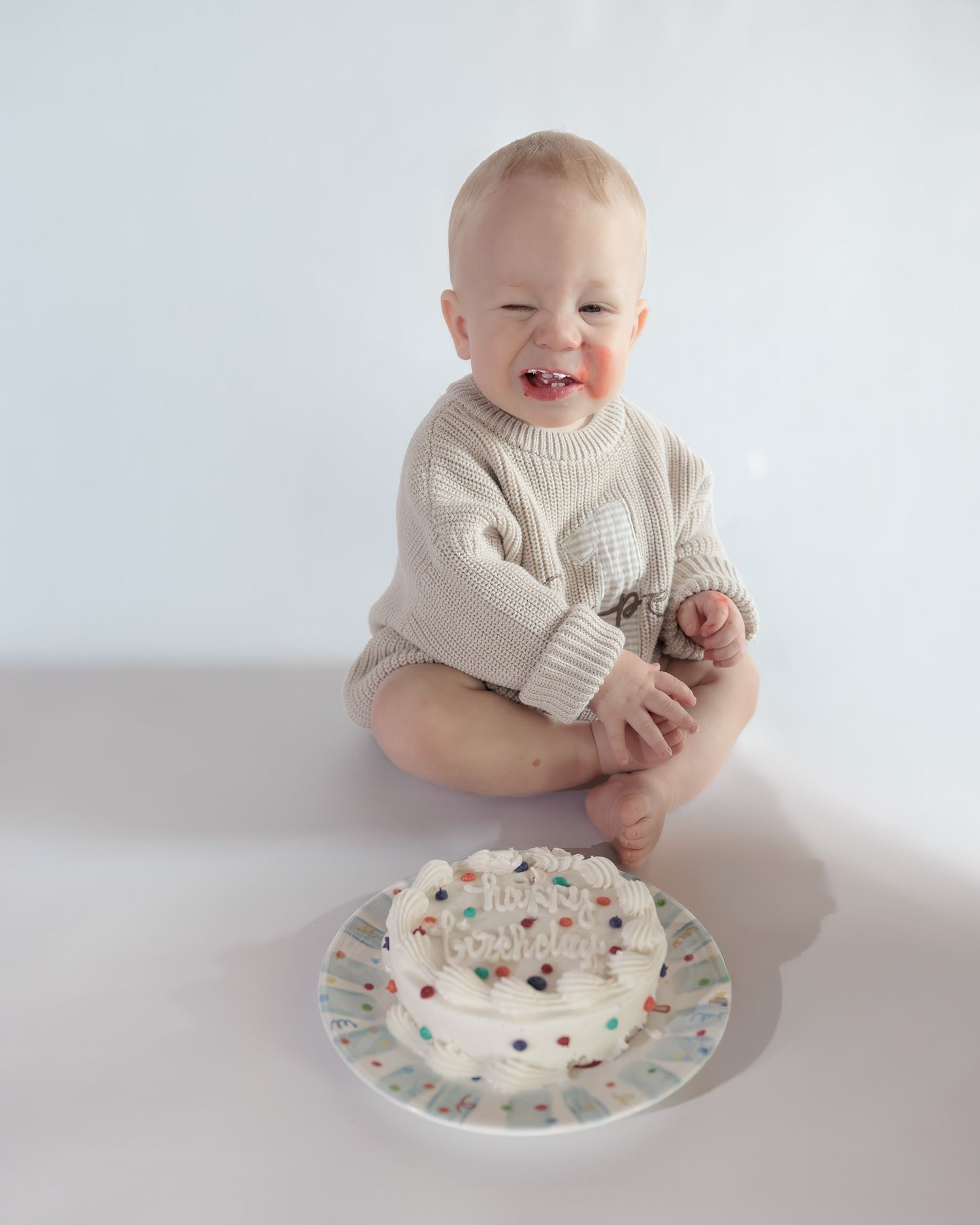 Smiling baby in a cream knit sweater sits beside a white sprinkle birthday cake on a white background.