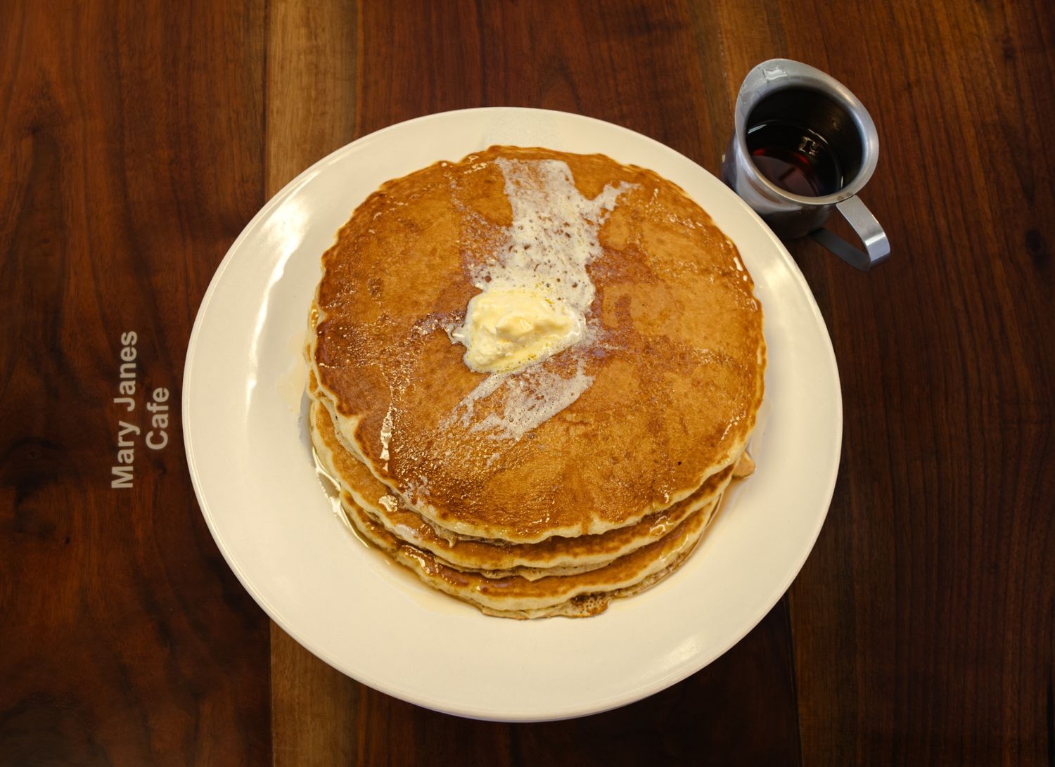 Stack of golden buttermilk pancakes topped with butter on white plate at Mary Janes Cafe with syrup cup.