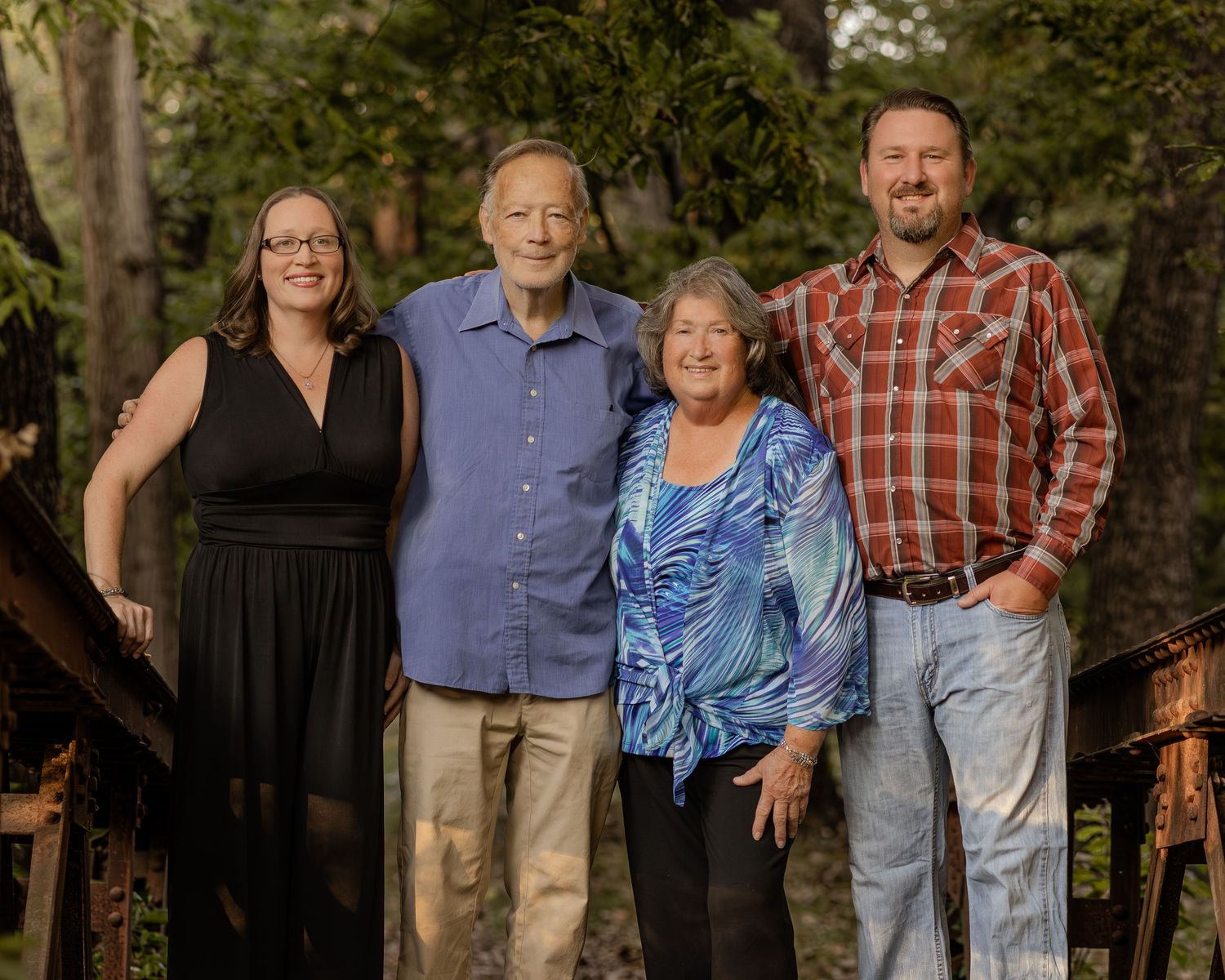 Four family members smiling together outdoors in a wooded setting, dressed in casual attire for a portrait photo.