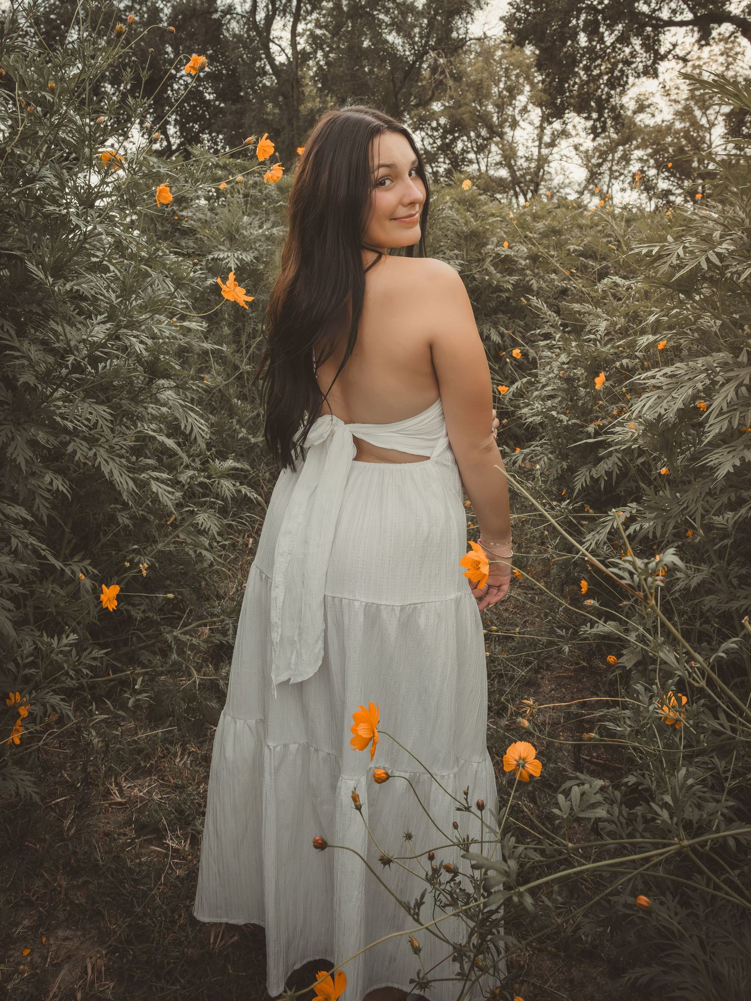 Brunette woman in white backless dress among orange flowers in garden.