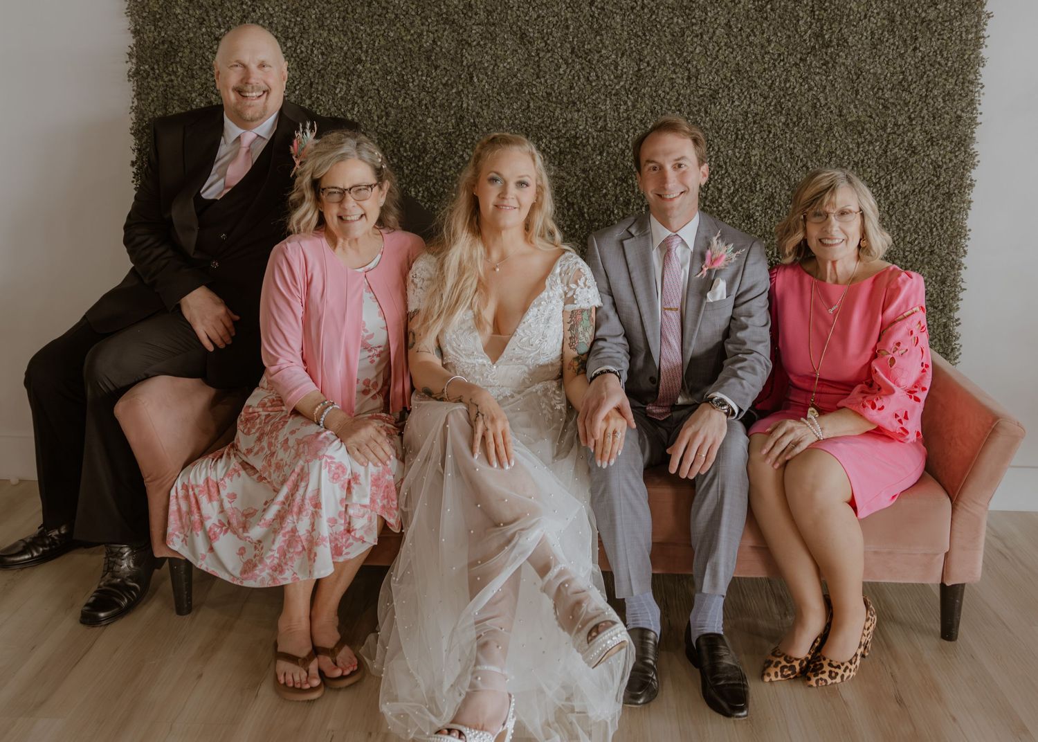Wedding party family portrait with bride in white gown and groom in gray suit seated on pink sofa.
